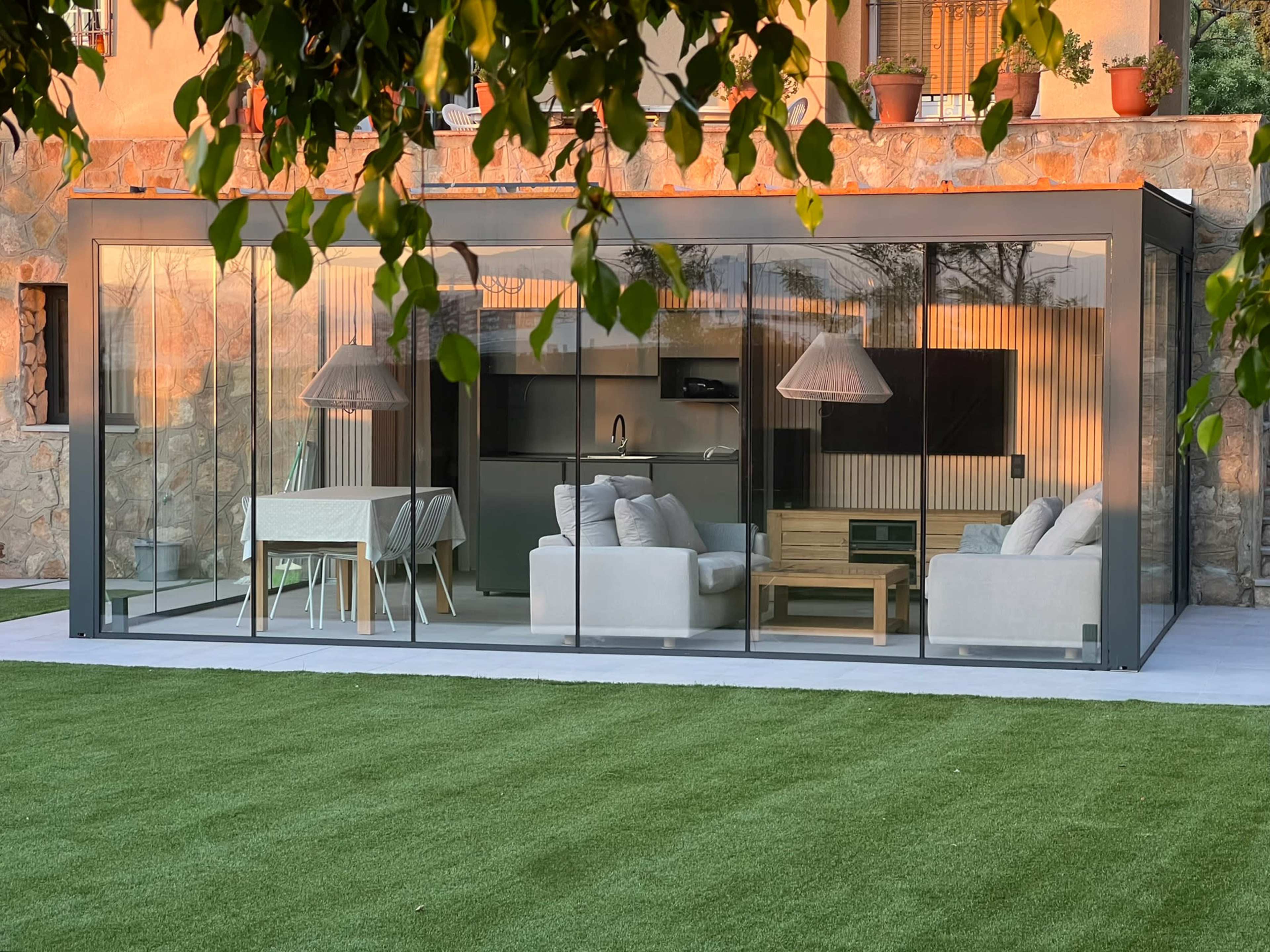 A modern glass-enclosed patio features a seating area with white sofas and a table, surrounded by artificial grass and stone walls.