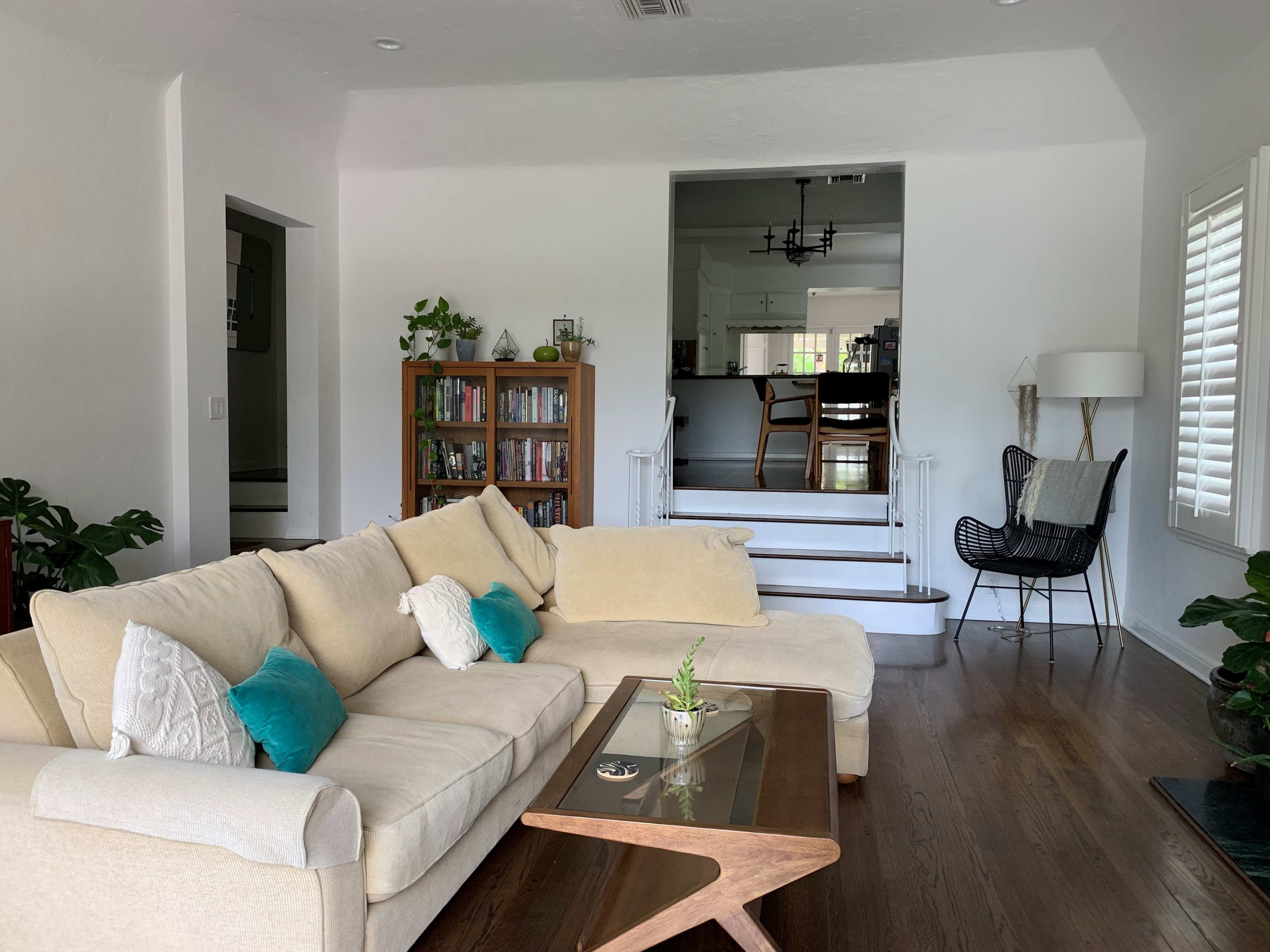 The image shows a living room with a beige sectional sofa, a glass-topped coffee table, and a bookshelf, leading to a dining area visible in the background.