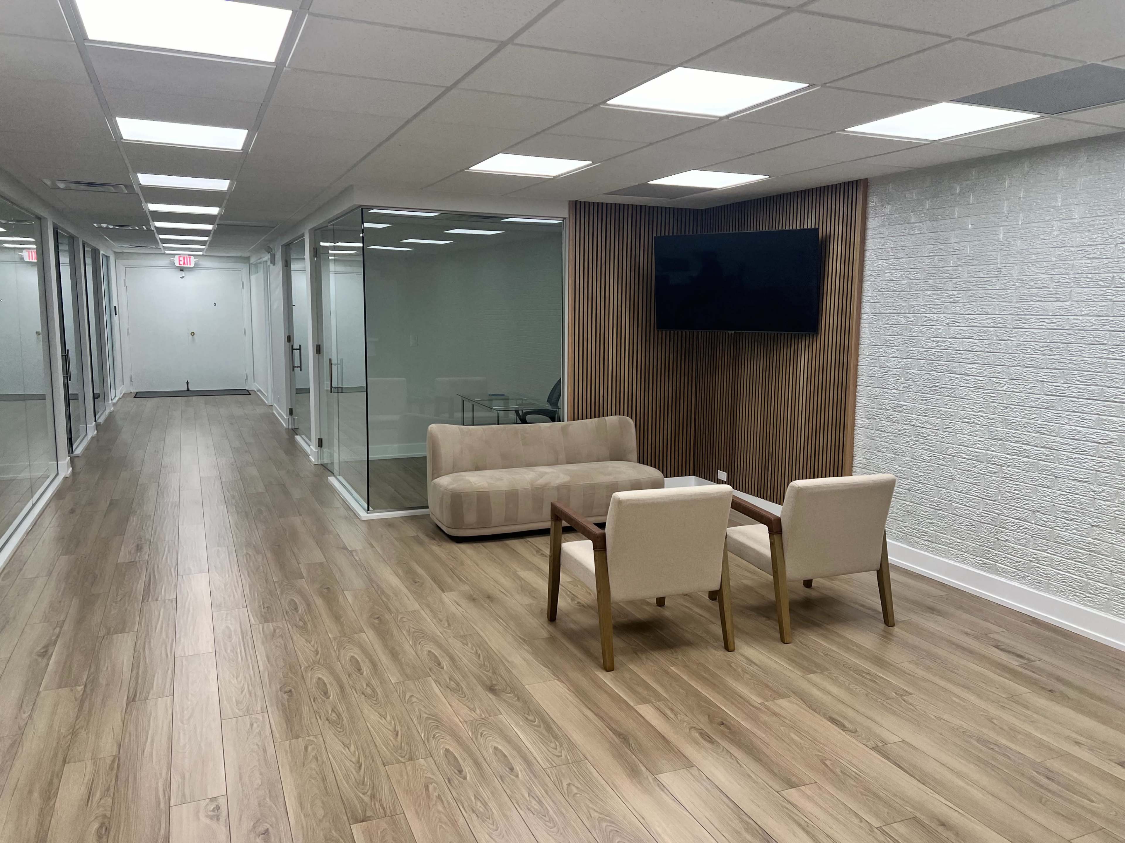 A modern office hallway with a seating area featuring a sofa and two chairs, alongside glass-walled conference rooms and a wall-mounted television.