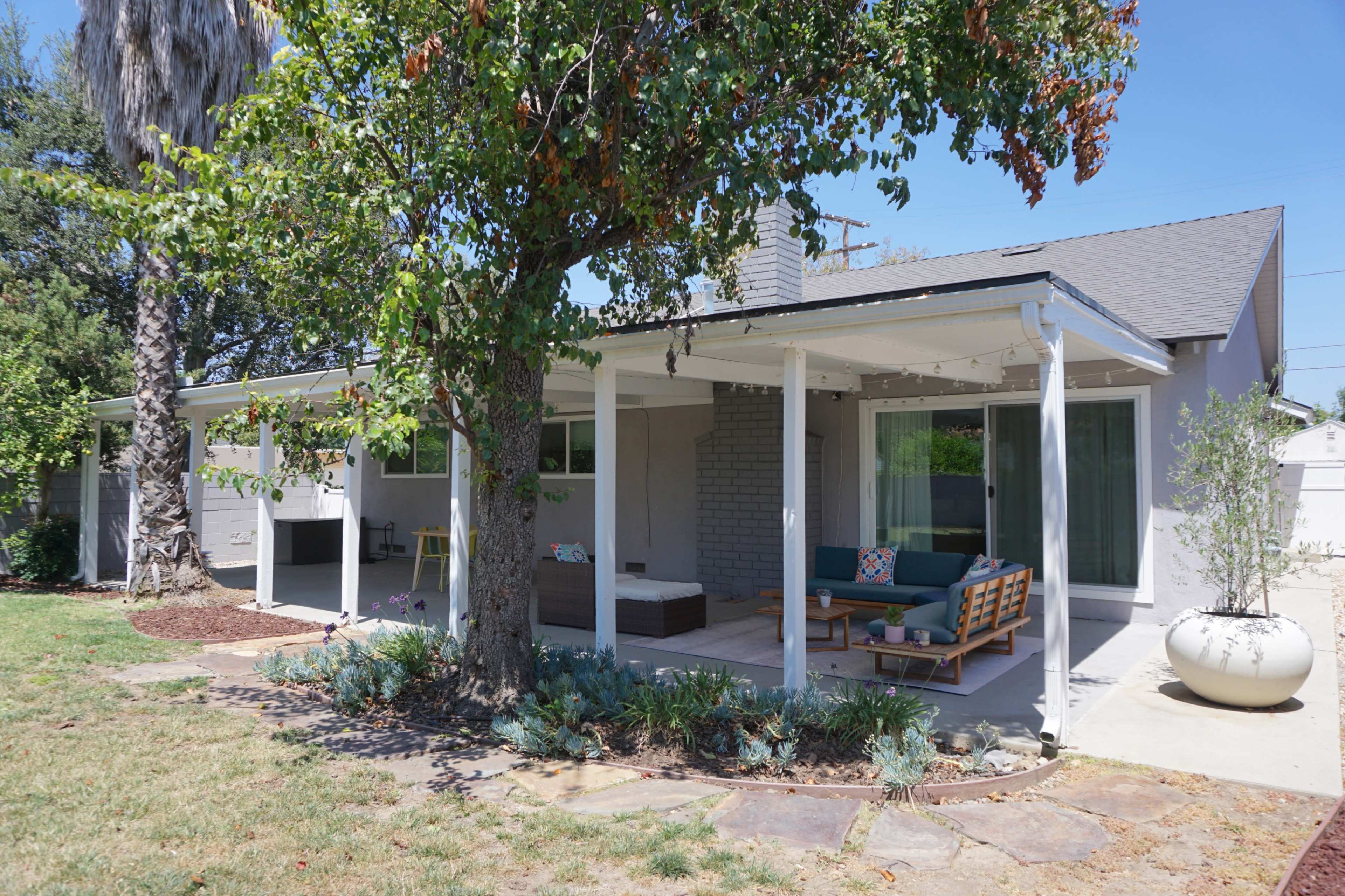 A single-story house with a covered porch features outdoor seating and greenery in the front yard.