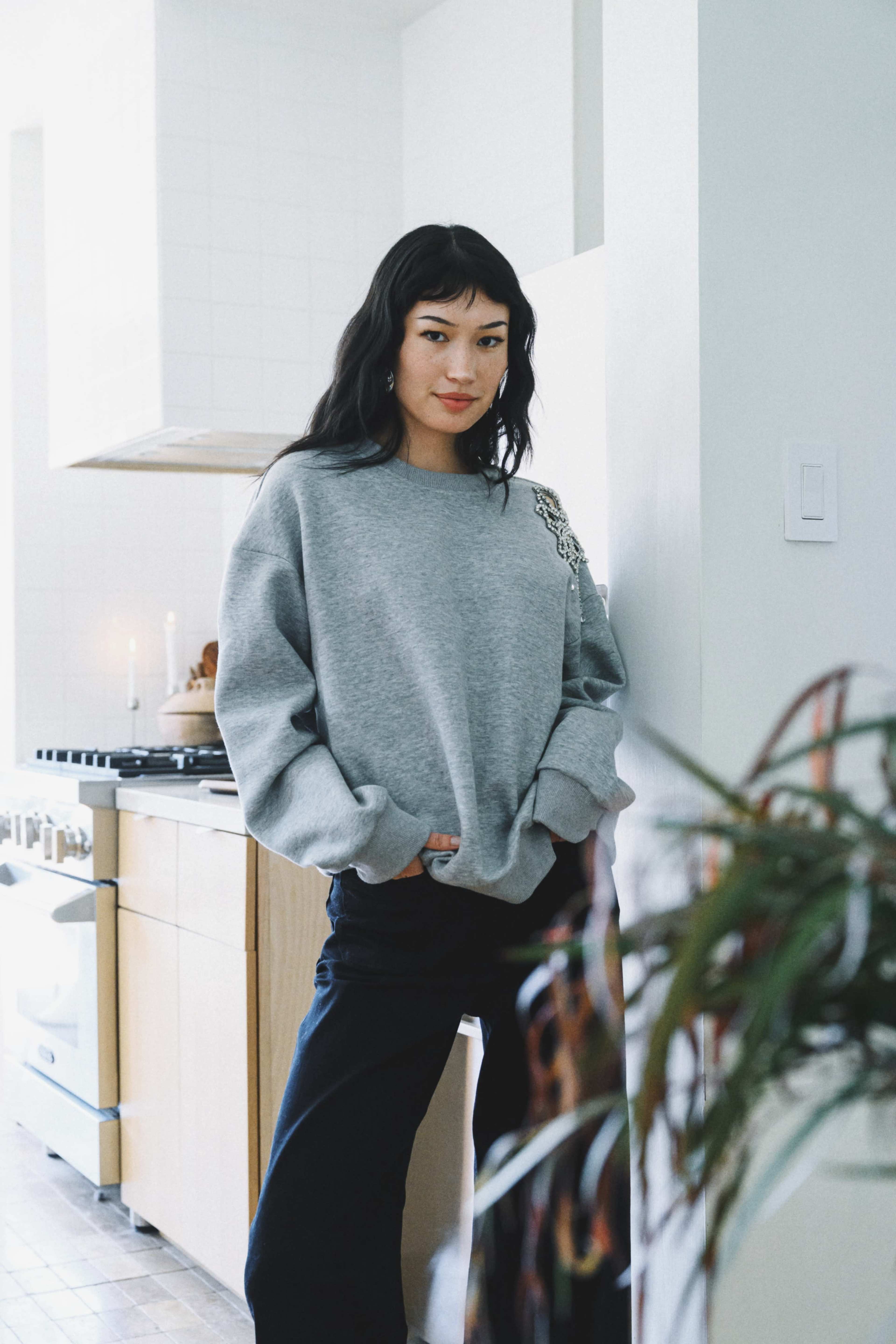 A woman in a gray sweatshirt stands against a kitchen wall with wooden cabinets and a stove in the background.