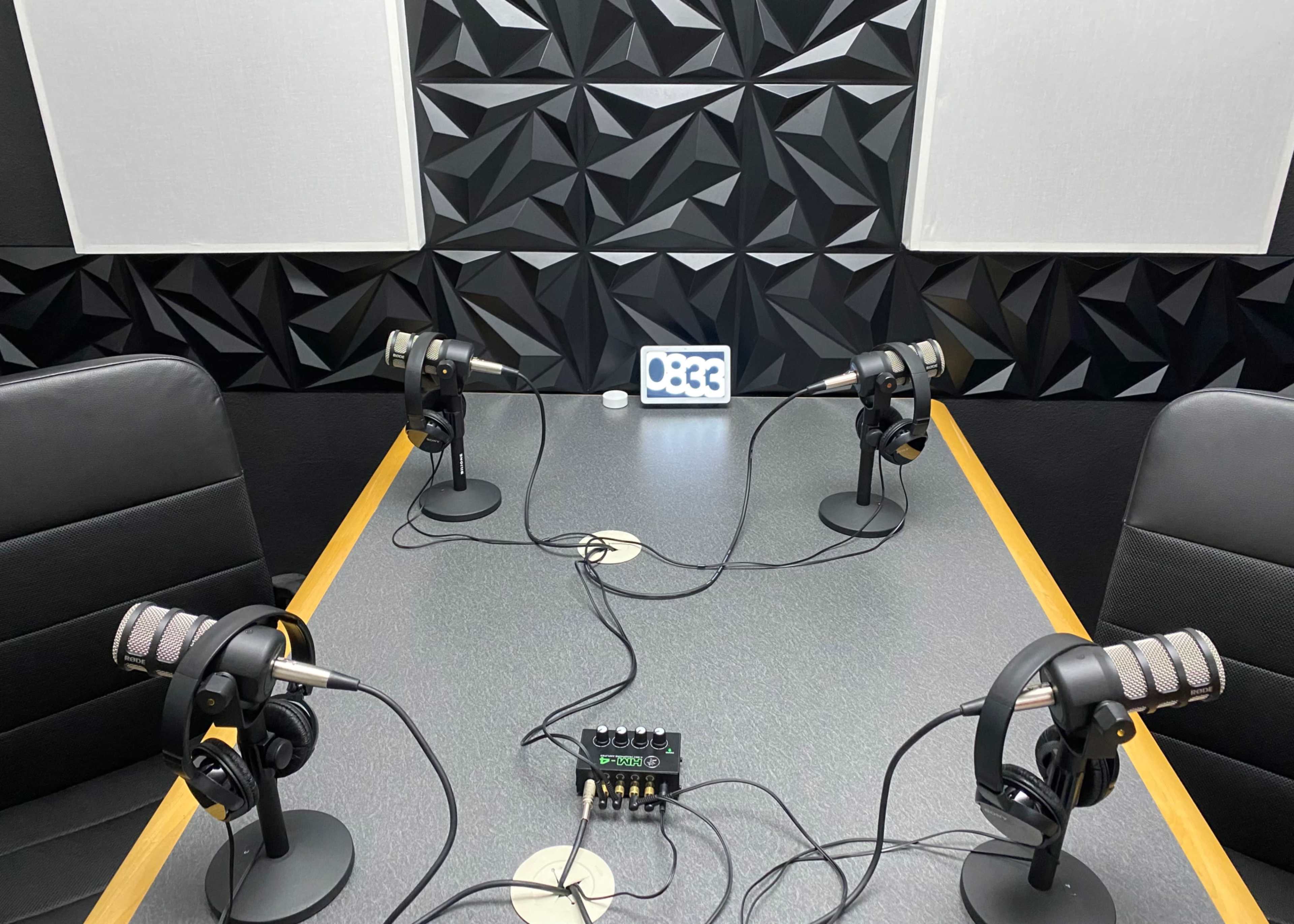 The image shows a podcast recording studio with four microphones, headphones, and a mixer on a table surrounded by textured black walls.
