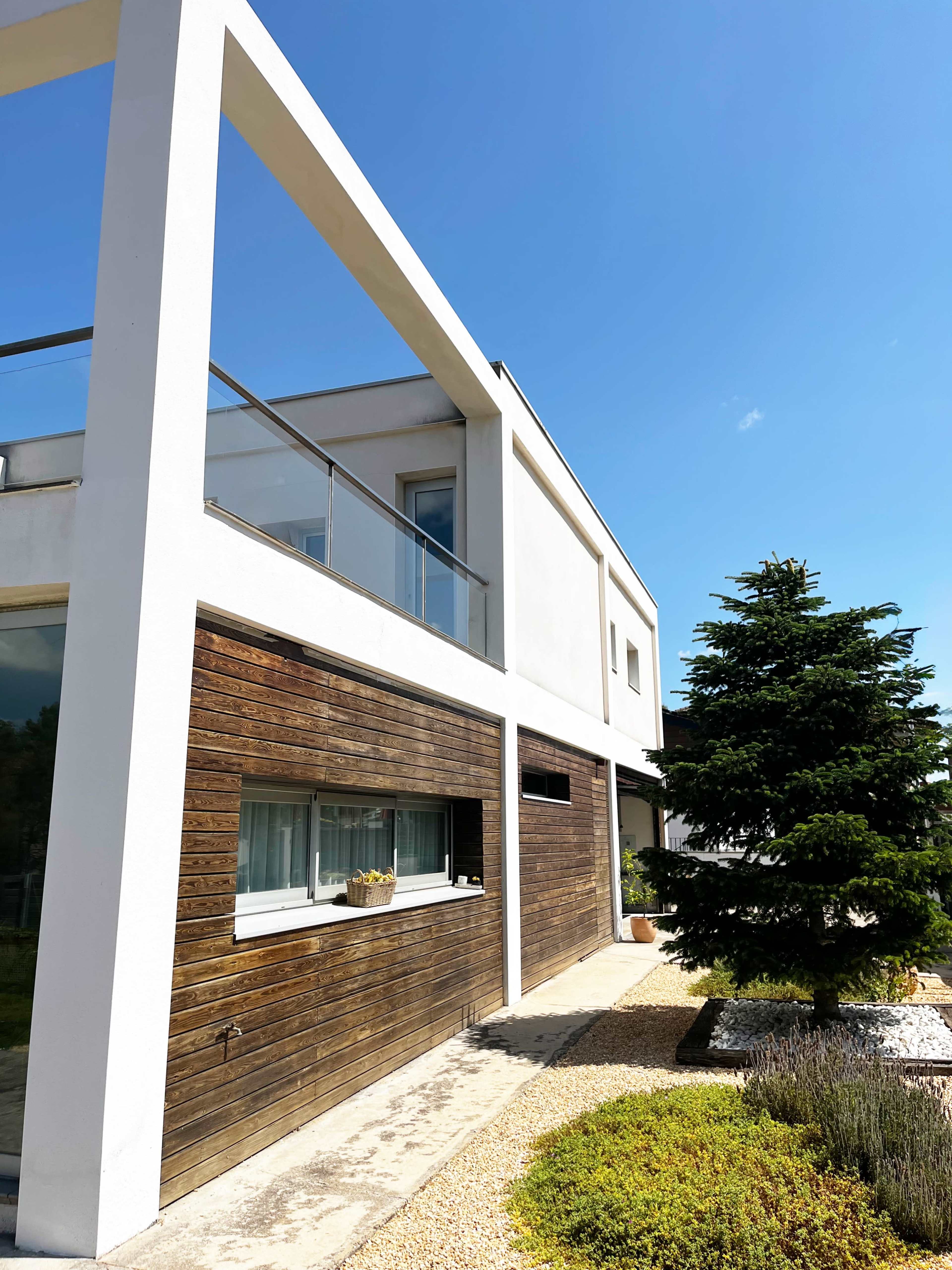 The image shows a modern house with a wooden façade, a large window, a balcony, and a well-maintained garden featuring a conifer tree.