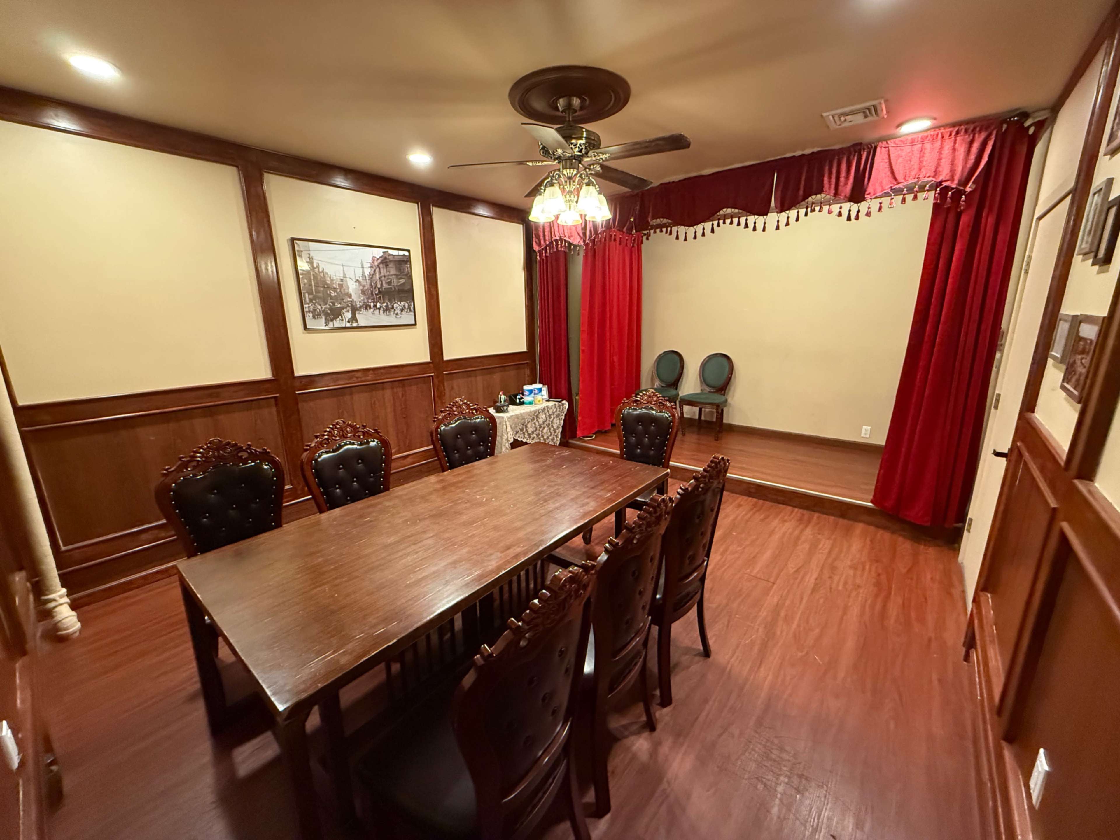 The image shows a dimly lit dining room with a large wooden table surrounded by eight ornate chairs, a set of two chairs on a small raised platform, and red curtains framing the stage area.