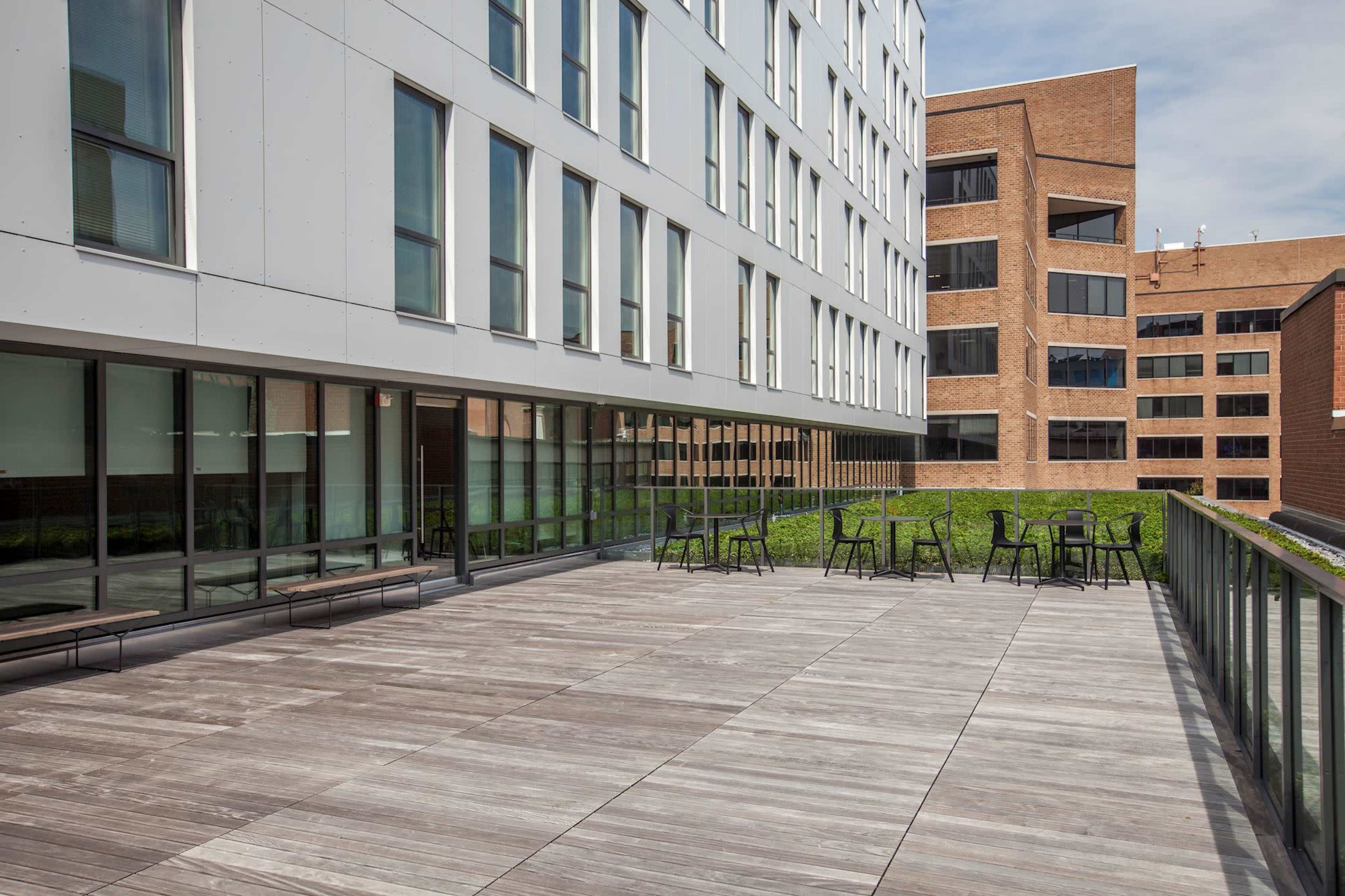 The image shows a spacious outdoor terrace with wooden flooring and metal furniture, surrounded by modern buildings.