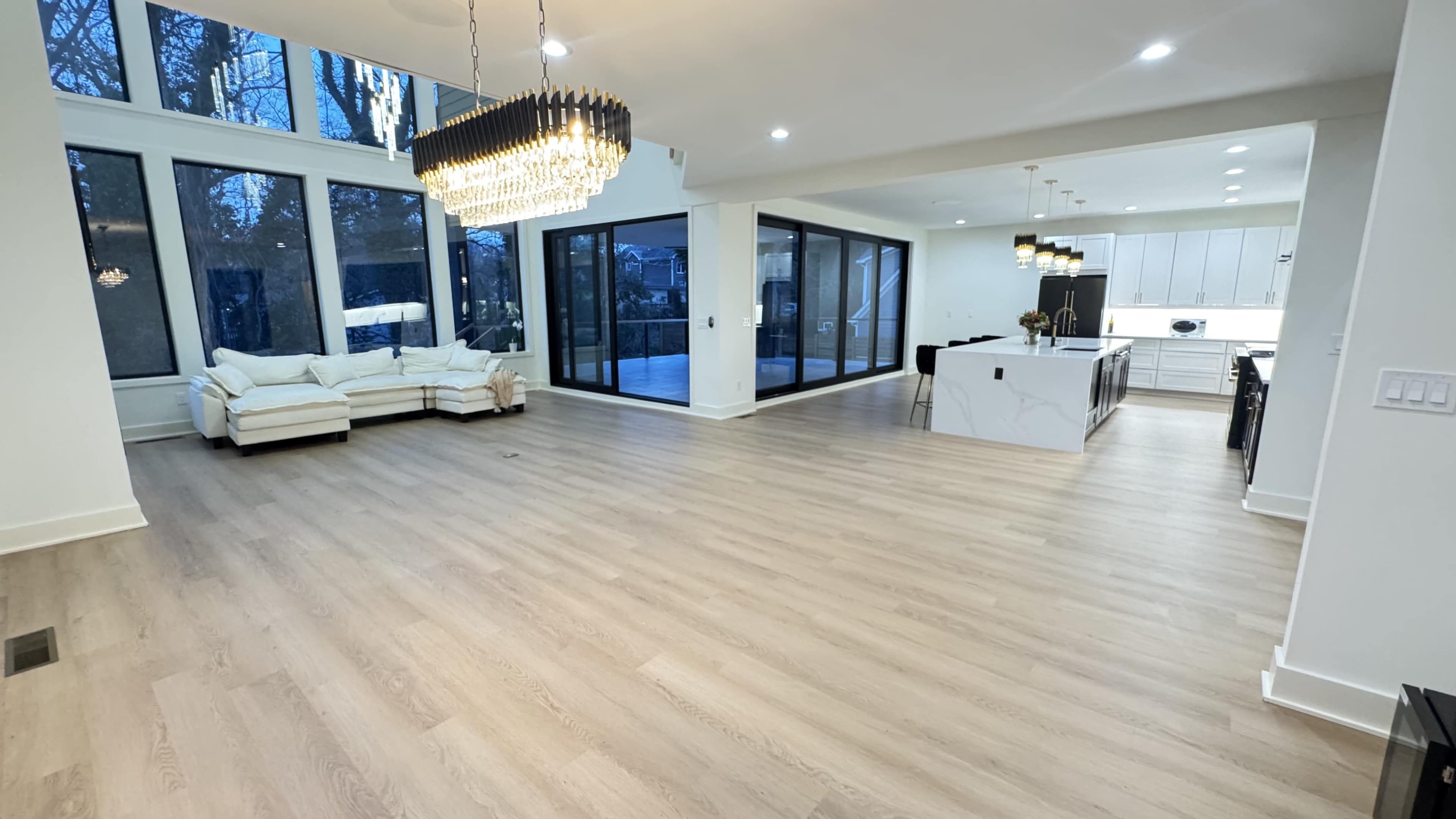The image shows a spacious, modern living area with large windows, a light-colored sofa, and a kitchen area featuring a marble island.