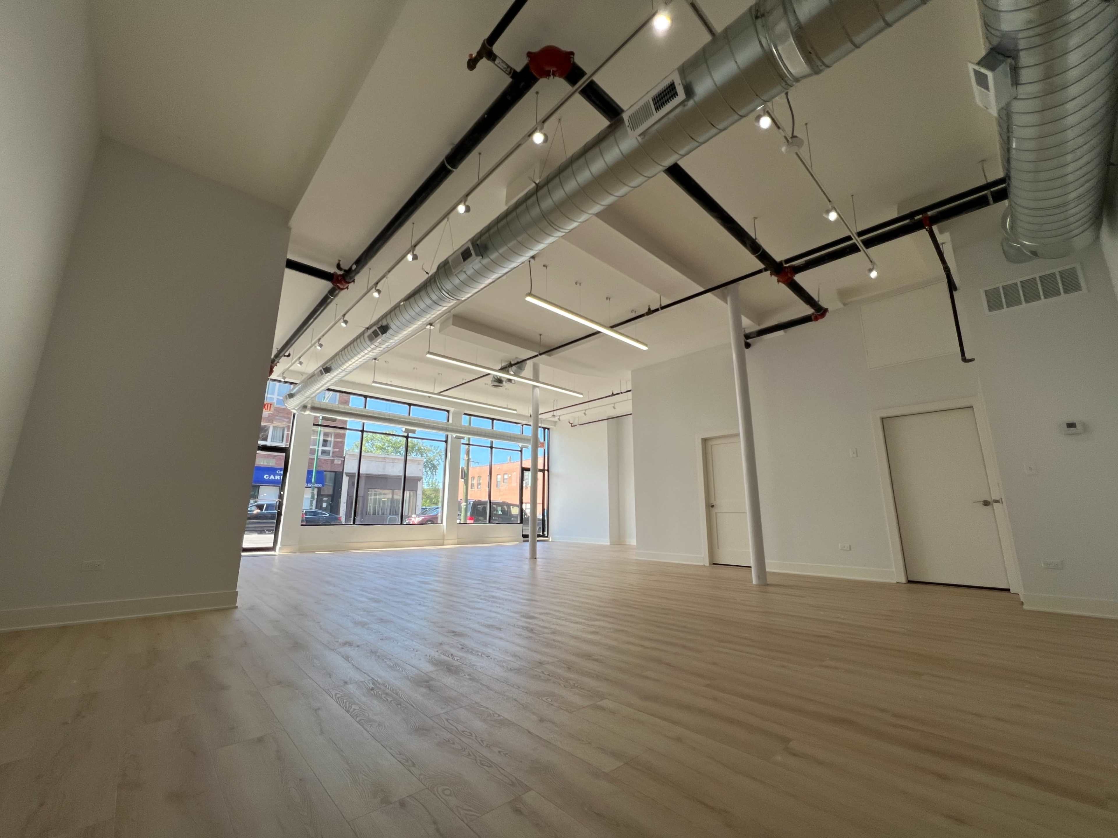 A spacious, empty room with wooden floors, large windows, and exposed ductwork on the ceiling.