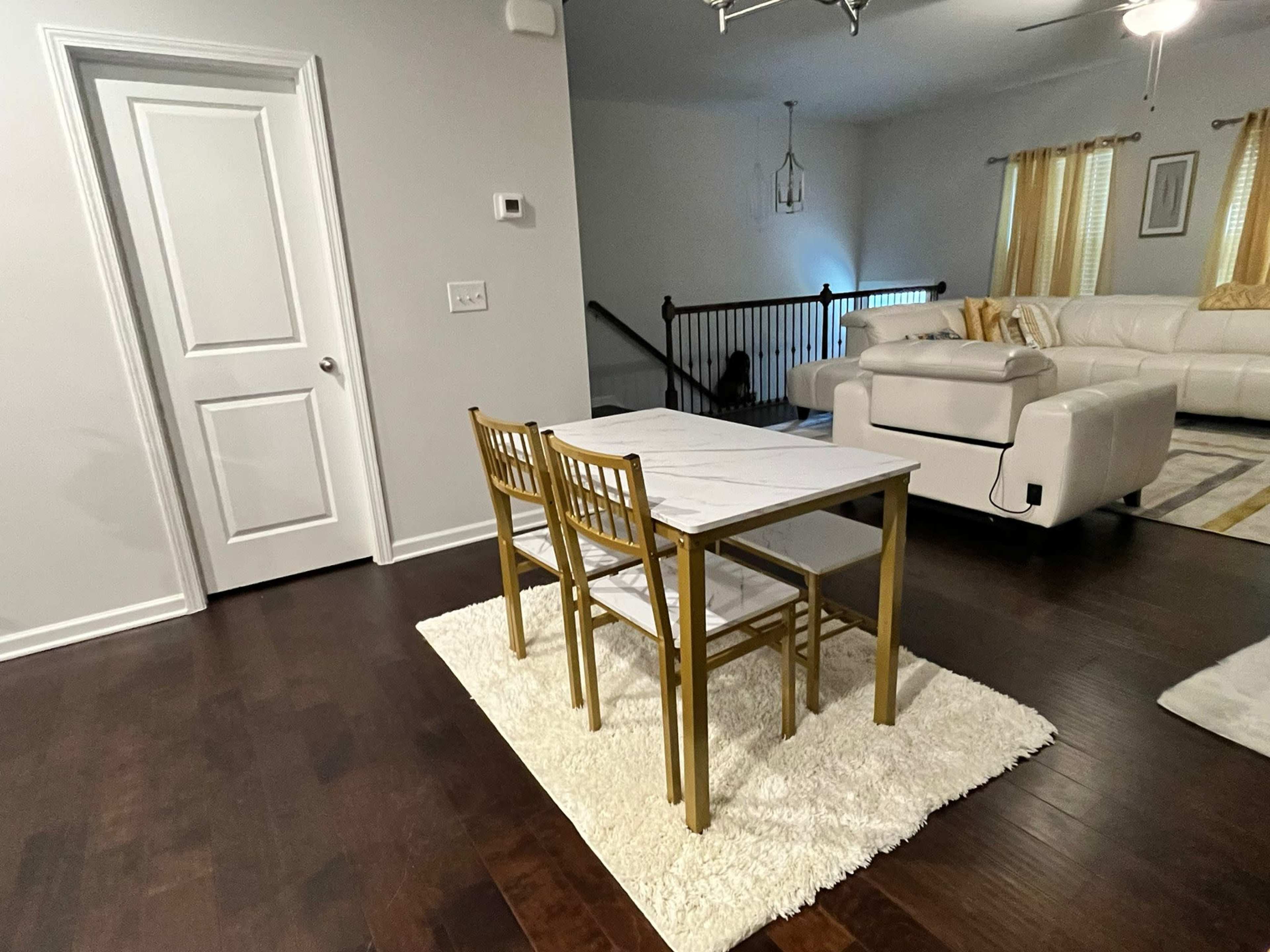 A small dining table with two chairs is set on a white rug in a well-lit room featuring hardwood floors and a doorway leading to another area.