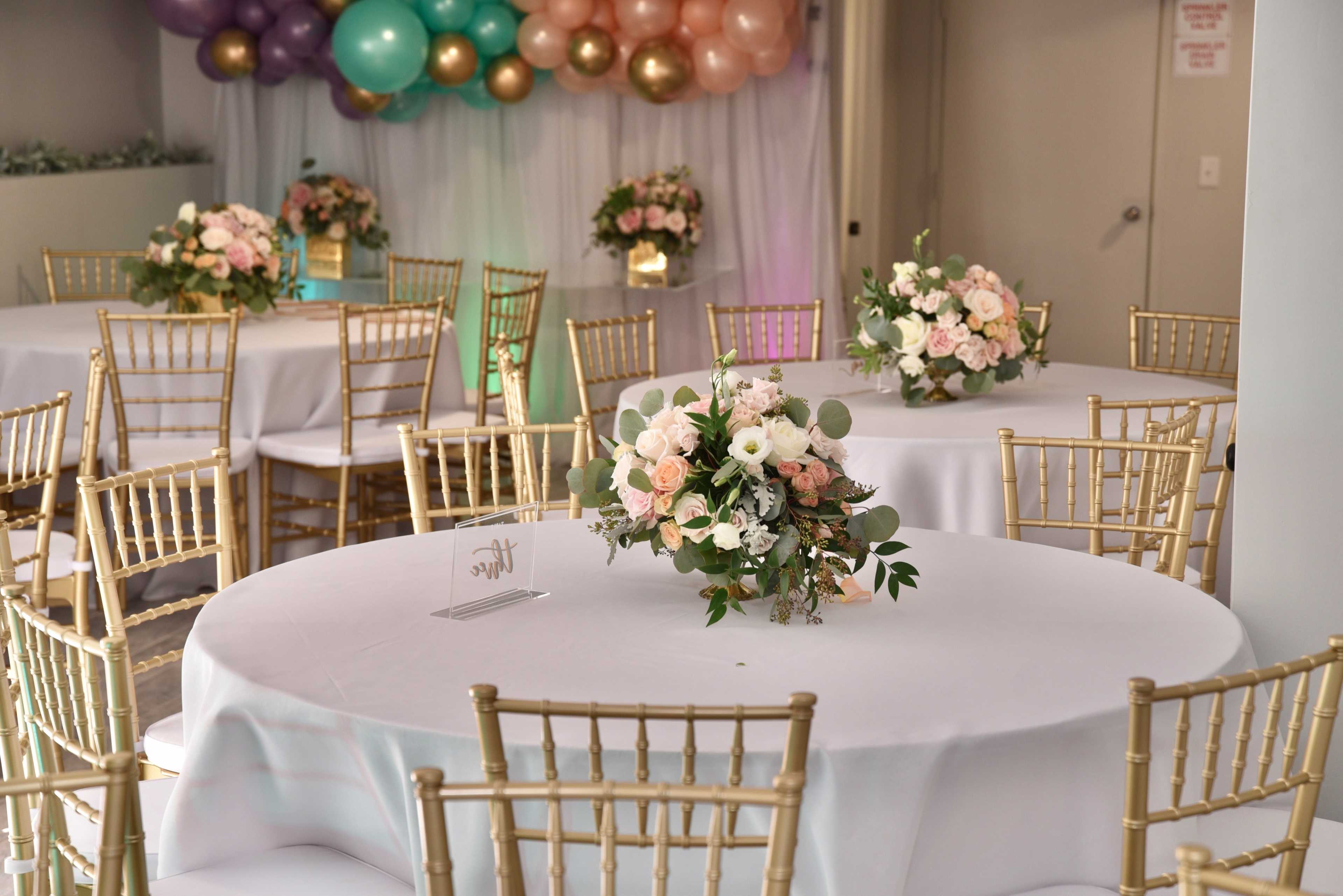 A banquet hall set up for an event, featuring round tables with white tablecloths, gold chairs, and floral centerpieces, along with a decorative balloon backdrop.