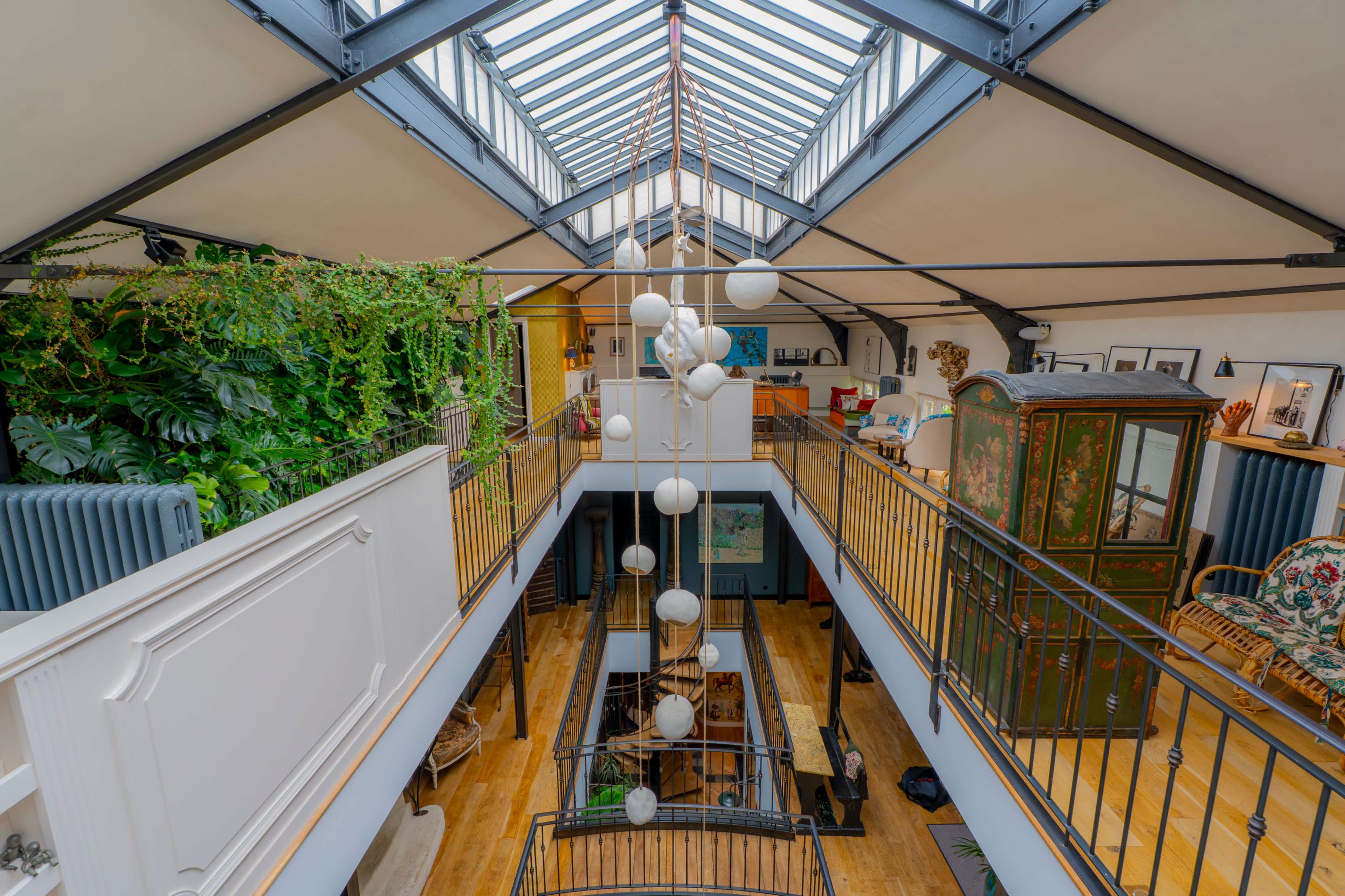 The image shows an open, spacious loft interior with a glass ceiling, plants cascading down, and a decorative arrangement of hanging lights.
