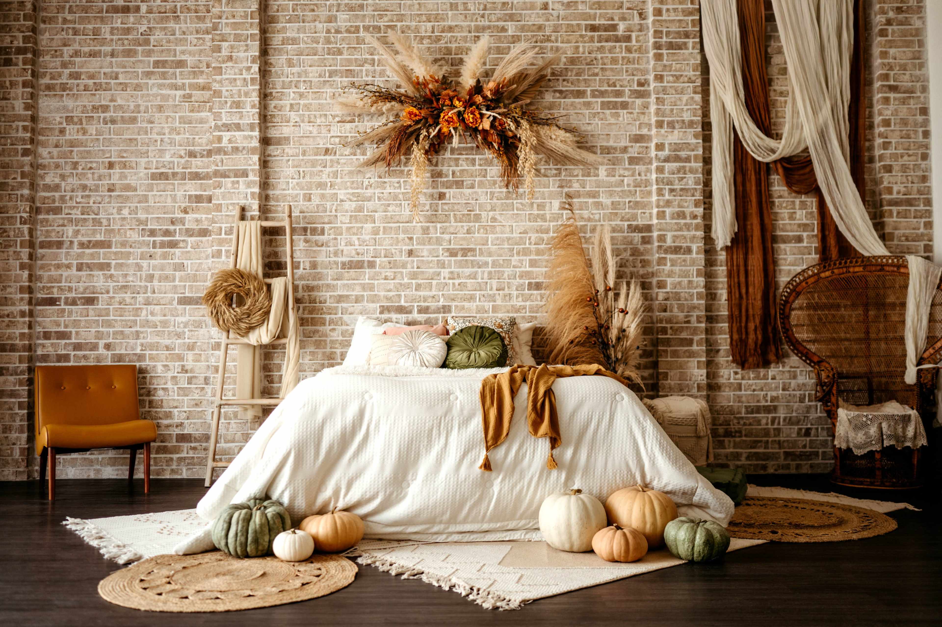 A cozy bedroom features a bed adorned with a white bedspread, surrounded by decorative pumpkins and arranged with natural elements on a textured brick wall.