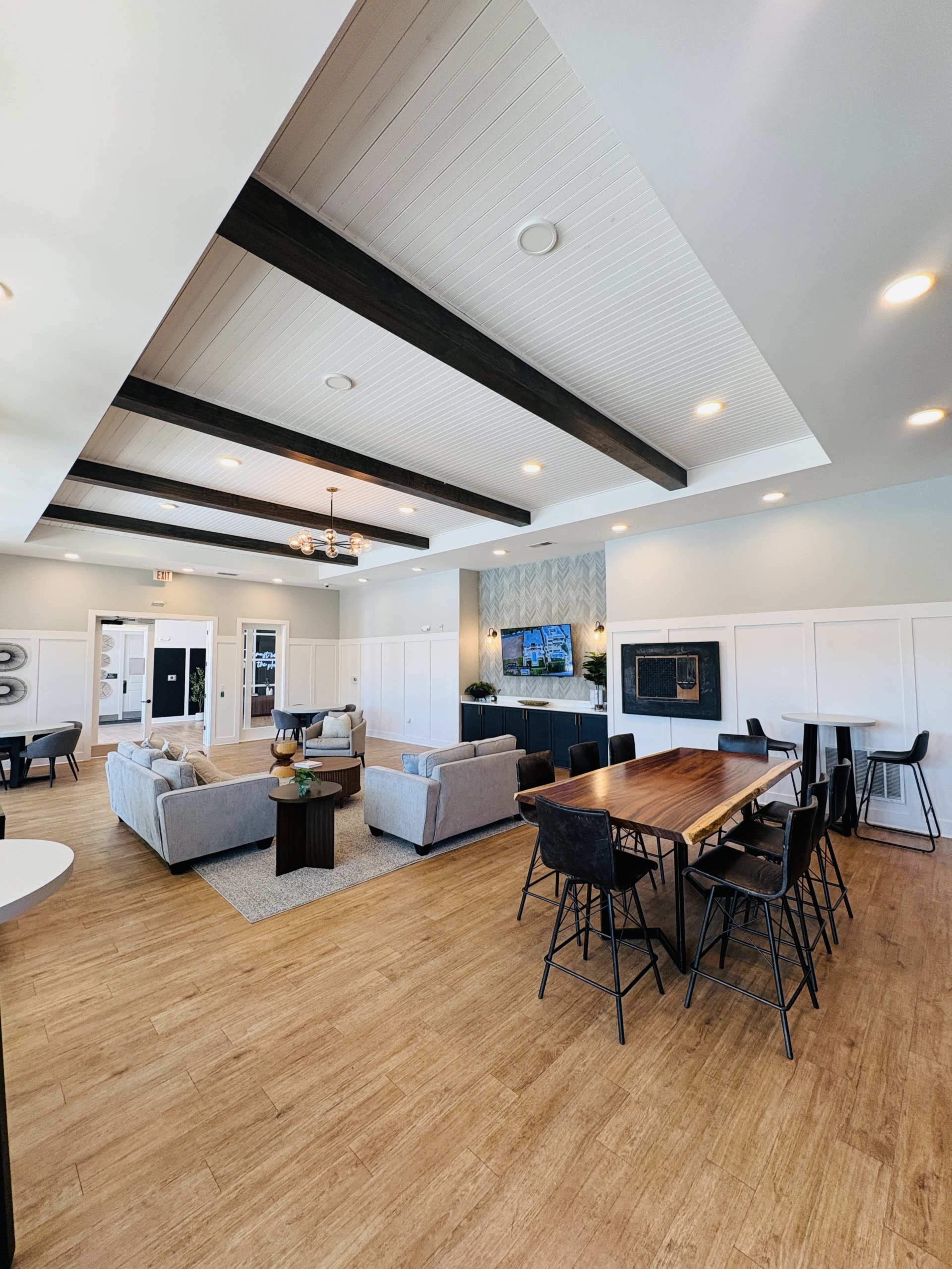 The image shows a modern communal lounge area with a seating arrangement of sofas and chairs, a dining table, and a wall-mounted television.
