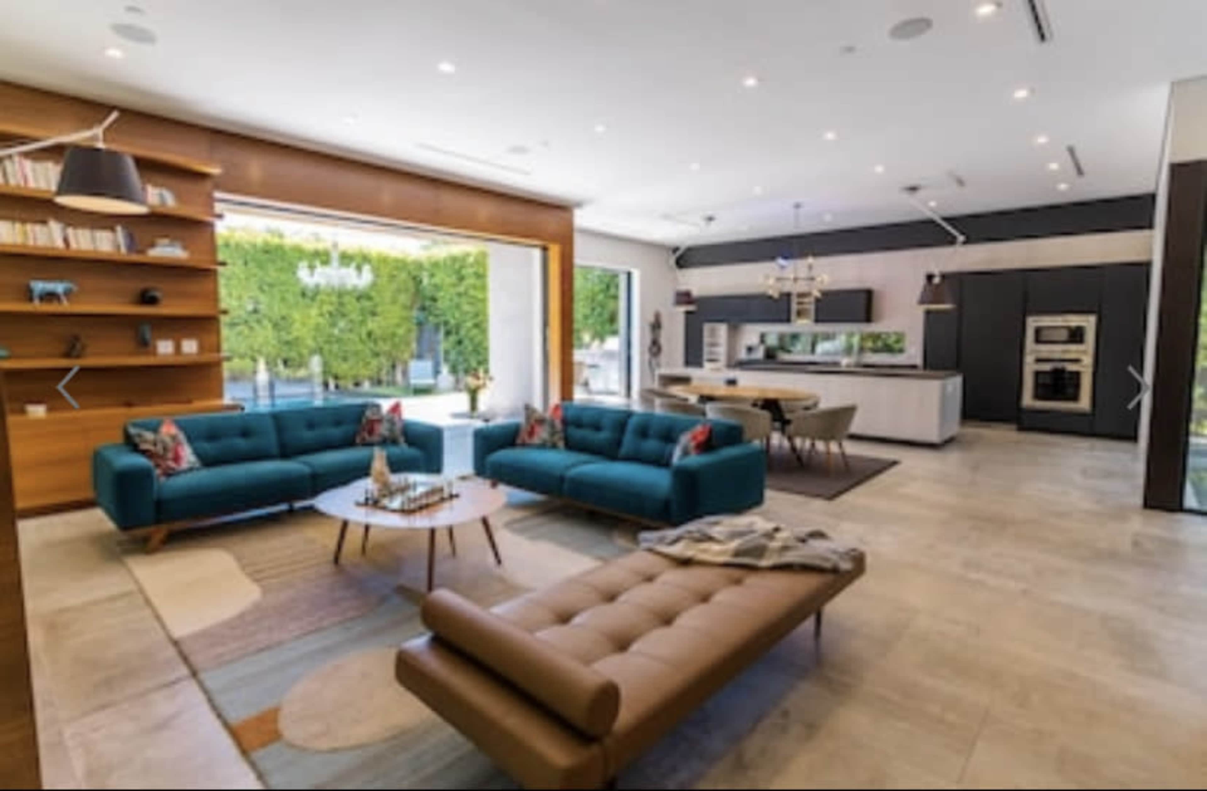 The image shows a modern living room with teal couches, a coffee table, and a dining area visible in an open layout, featuring large windows that lead to an outdoor space.