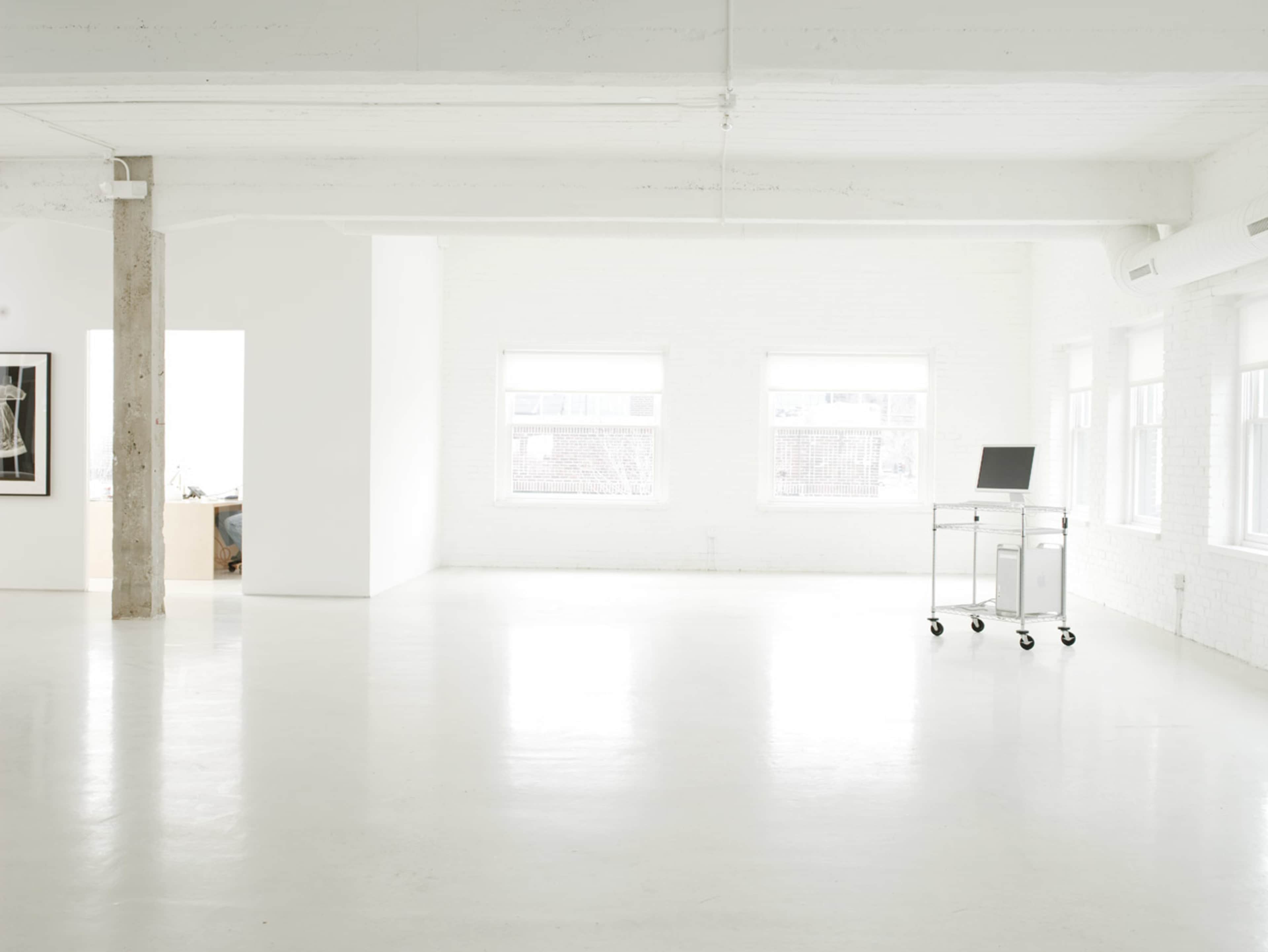 The image shows a spacious, minimalist white room with large windows, a concrete pillar, and a wheeled metal cart bearing a laptop.