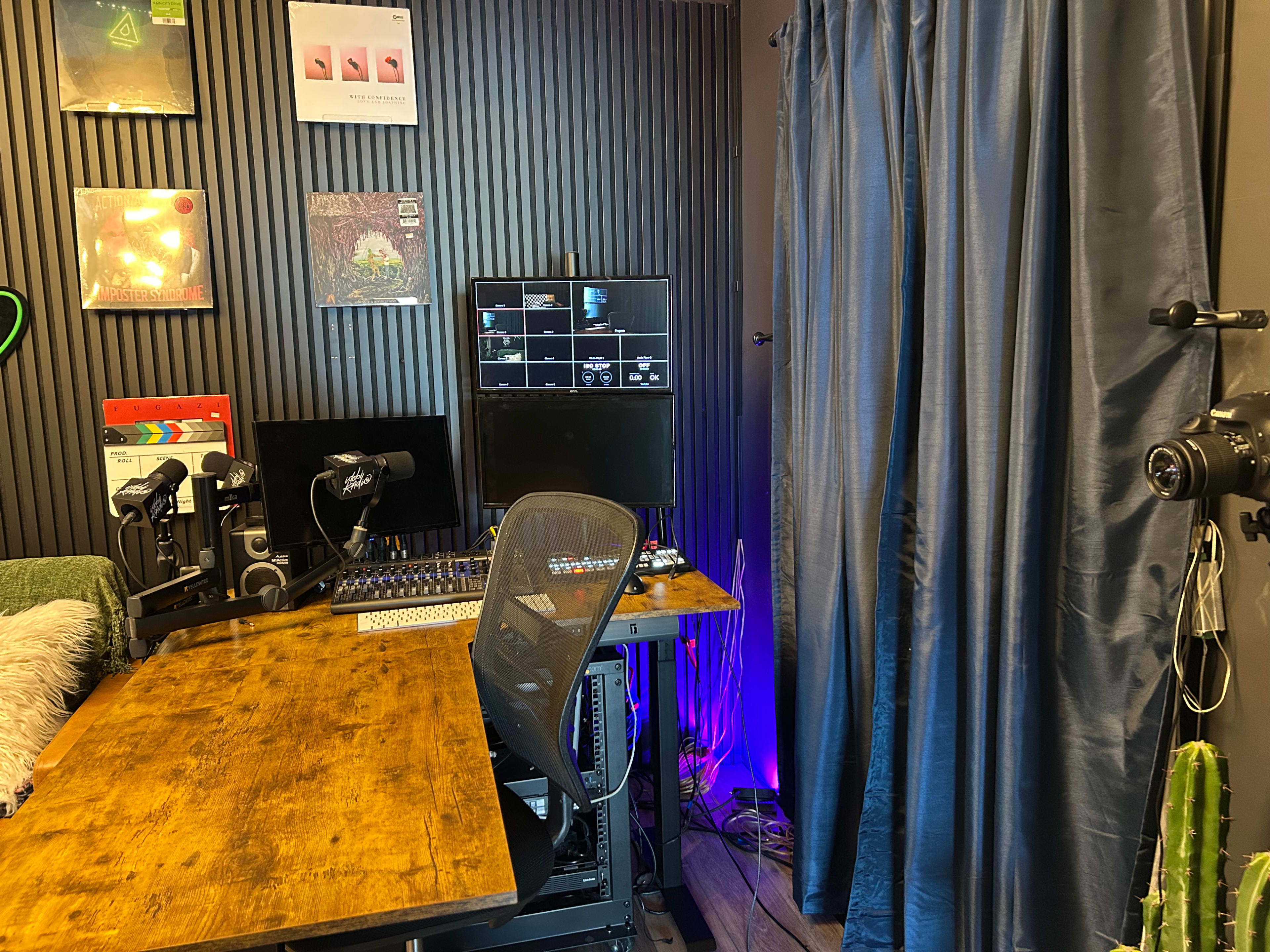 The image shows a small audio recording studio with a wooden desk, computer monitor, microphones, and a curtain on one side.