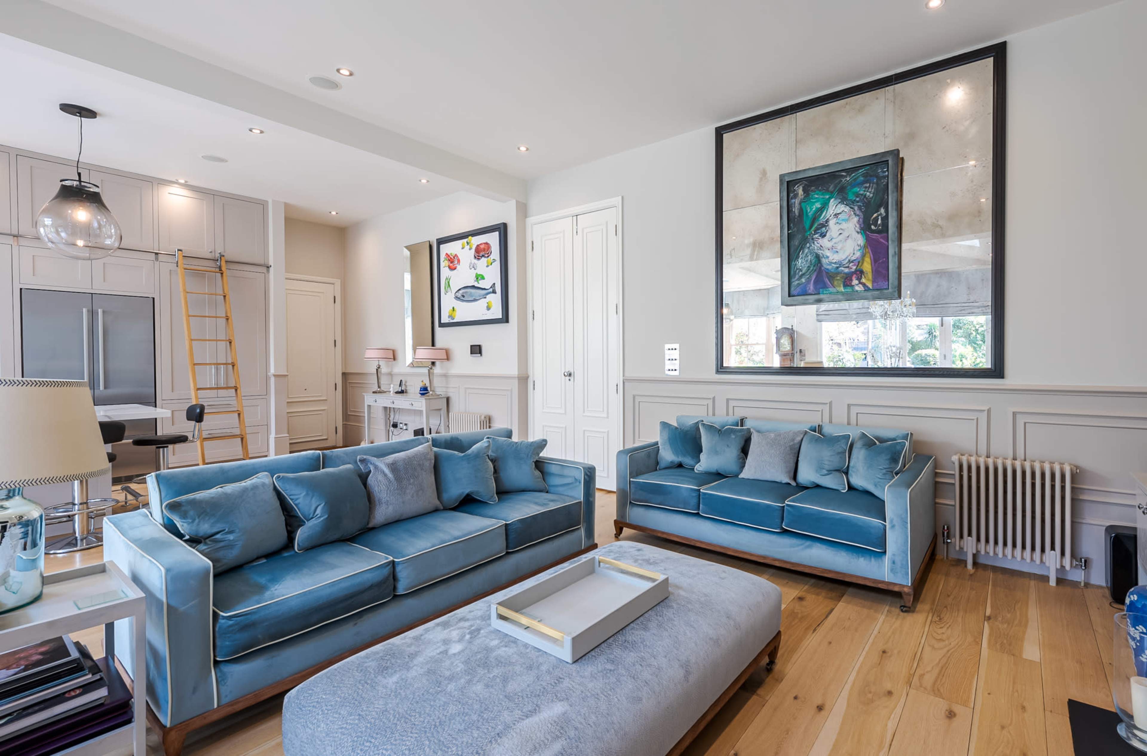 The living room features two blue velvet sofas facing a coffee table, with artwork displayed on the walls and light wood flooring.