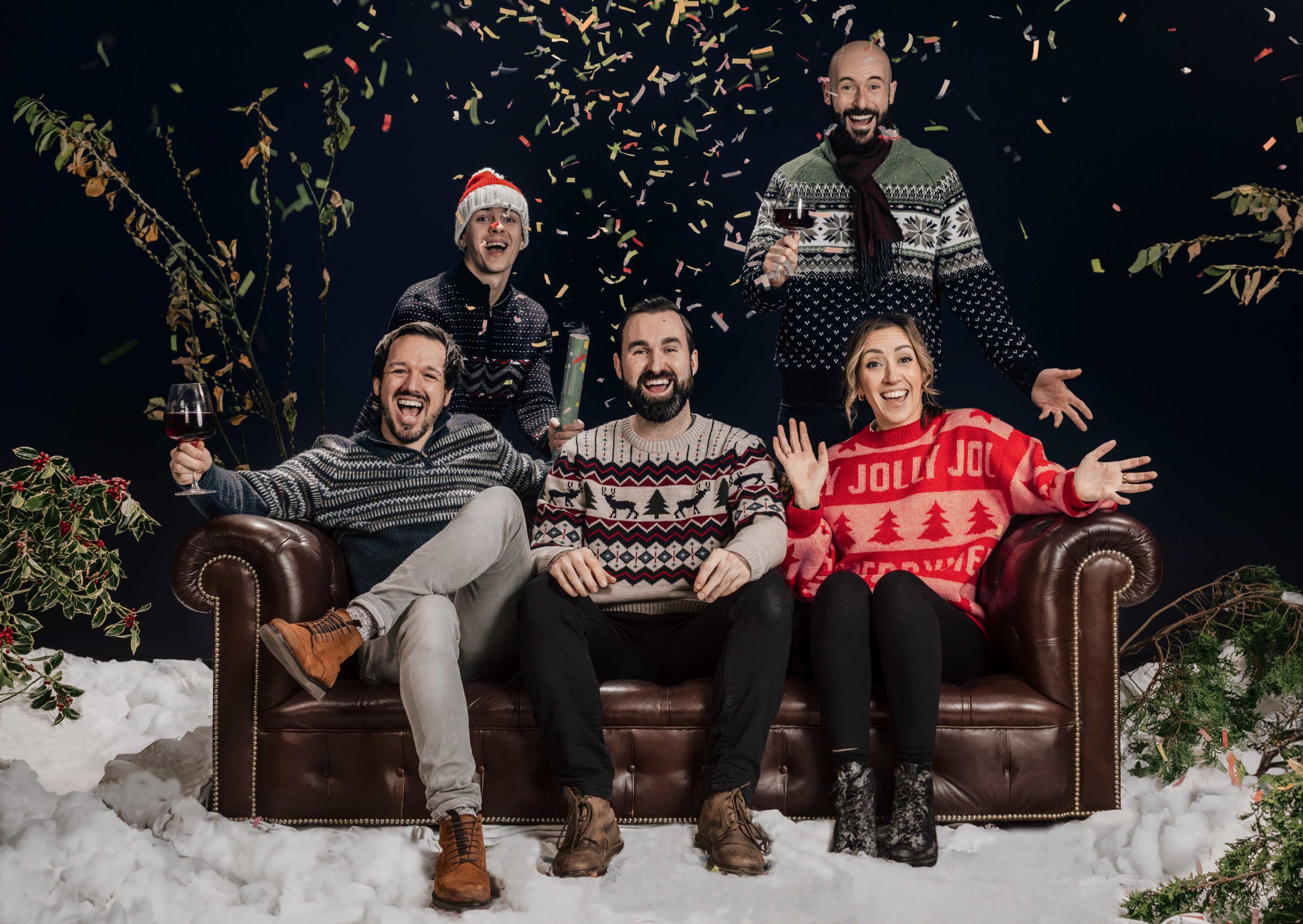 Five people wearing festive sweaters celebrate on a couch with confetti falling around them in a winter-themed setting.