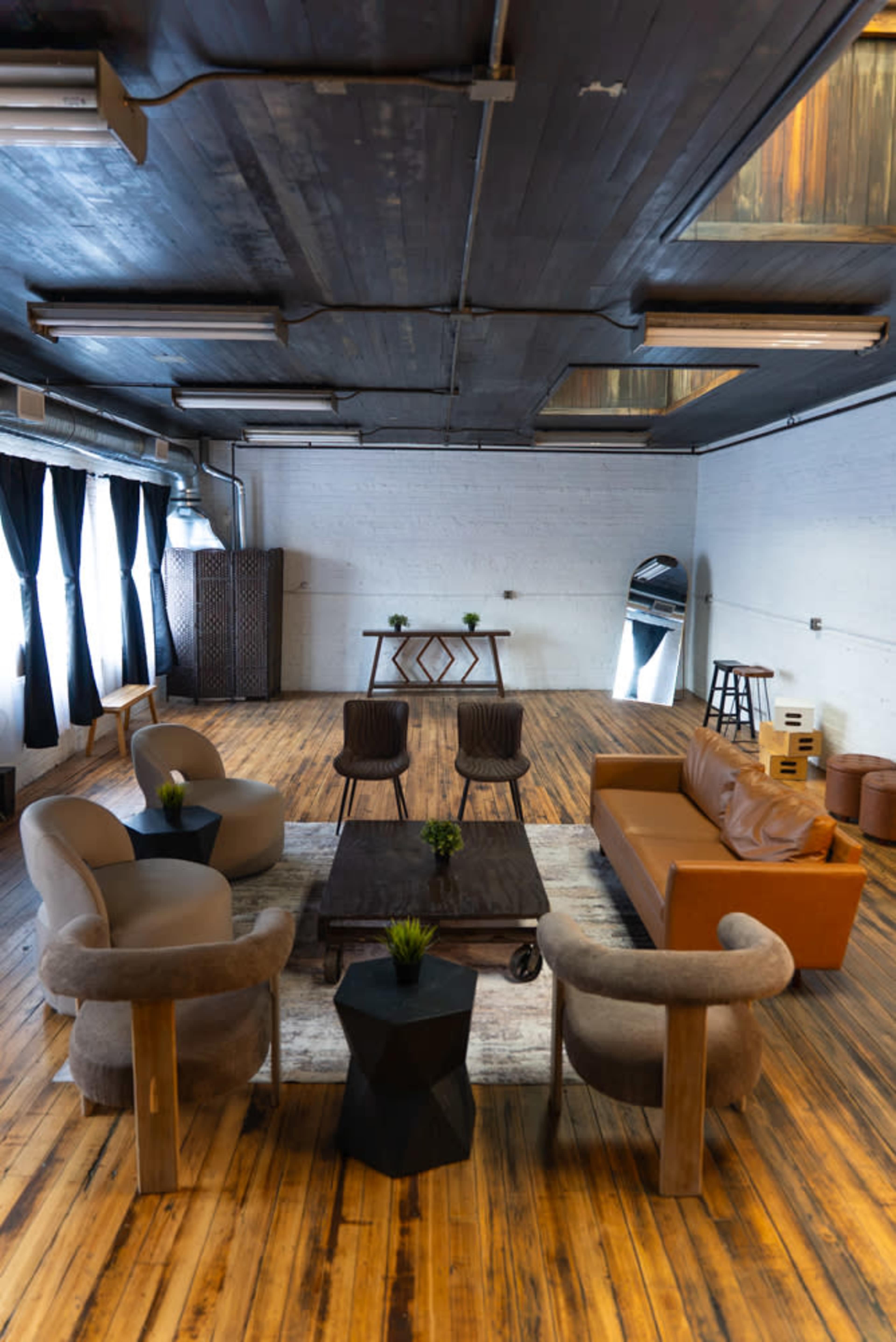 The image shows a spacious indoor lounge area featuring a mix of modern seating arrangements, a wooden table, and decor against a backdrop of wooden flooring and large windows.