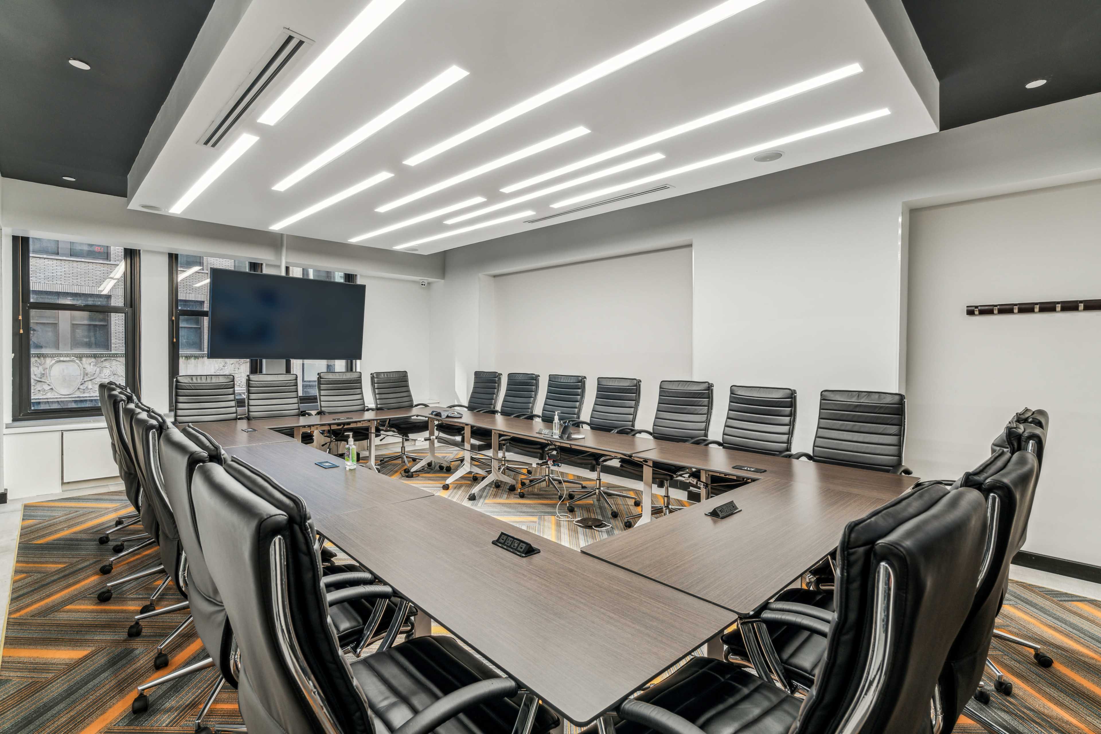 A modern conference room with a large rectangular table surrounded by black leather office chairs and a wall-mounted screen.