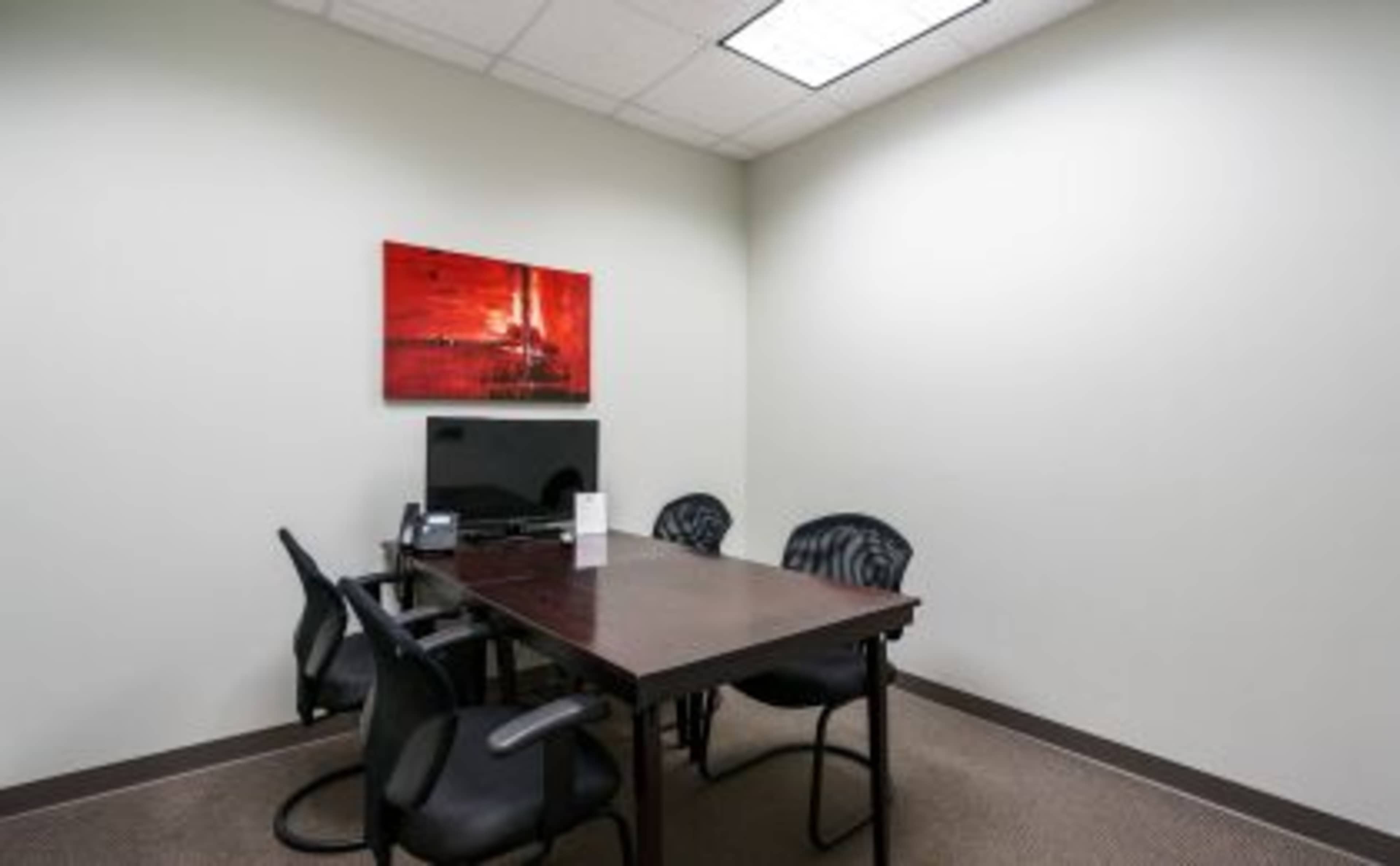 A small office room with a rectangular table, four black chairs, a television, and a piece of artwork on the wall.