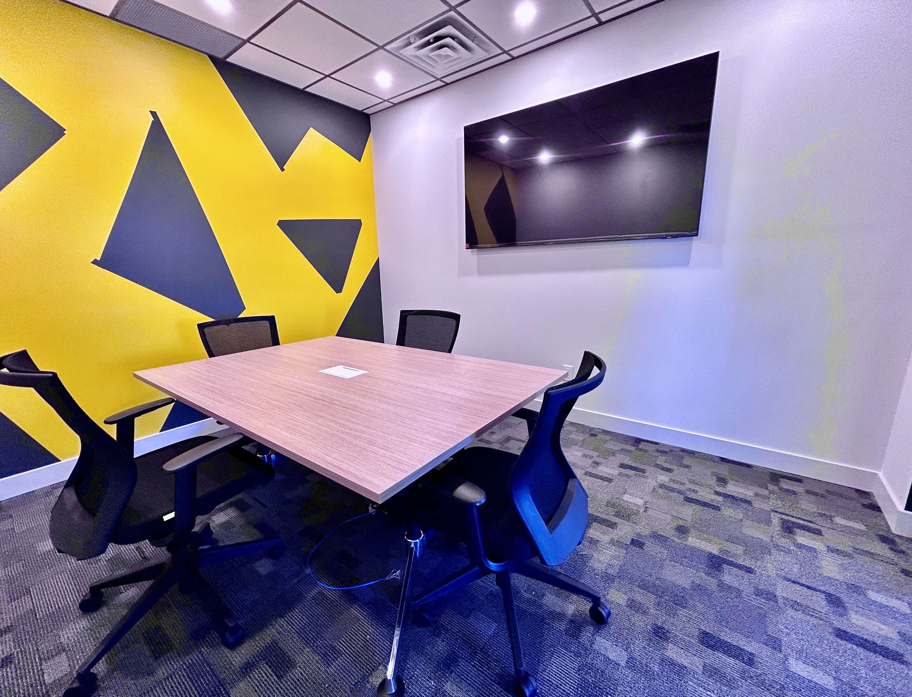 A meeting room features a wooden table surrounded by four black chairs, with a large television mounted on the wall and a yellow and gray geometric design on the backdrop.