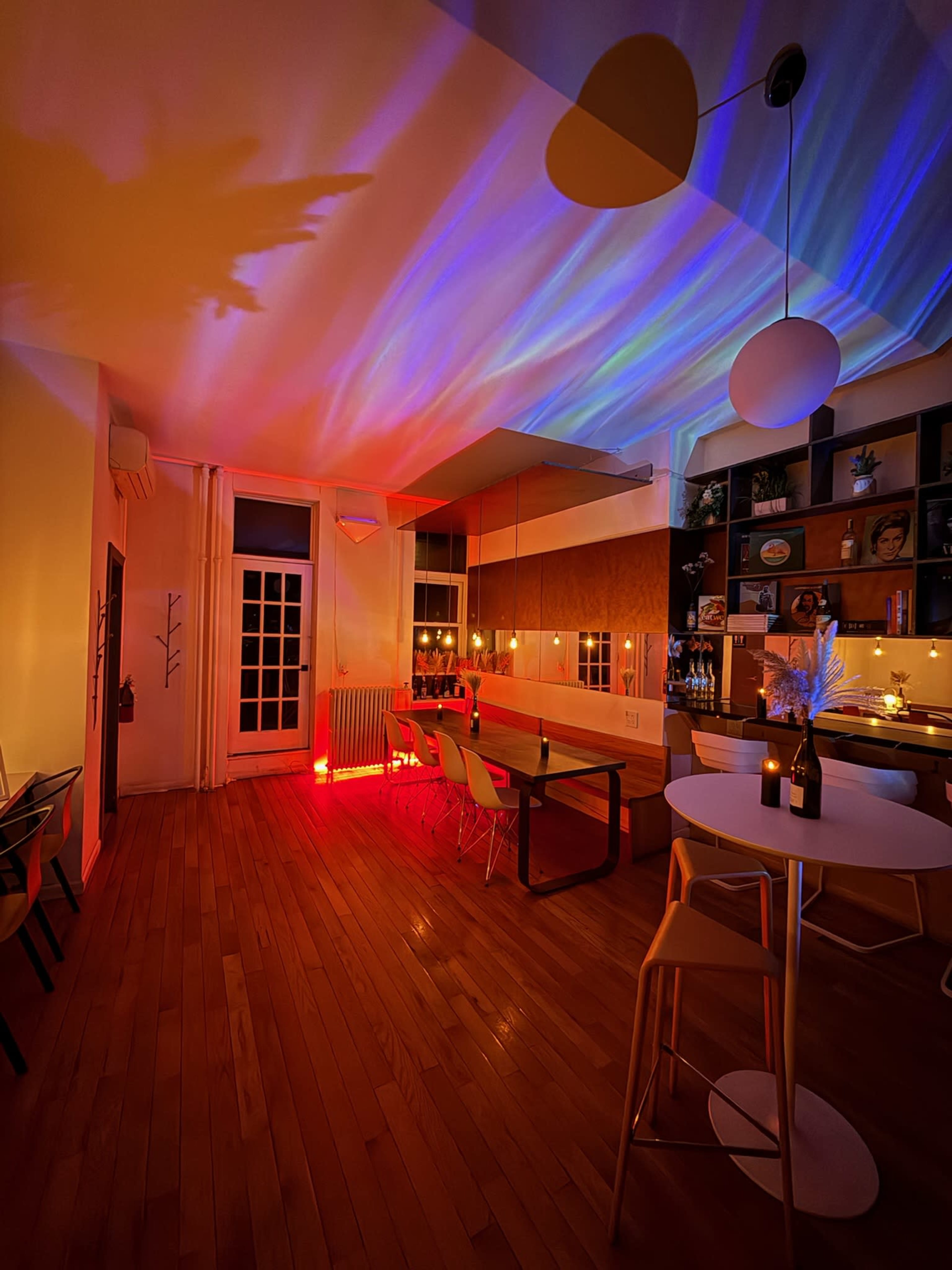 The image shows a modern dining area with a wooden floor, colorful light projections on the ceiling, and a table set with chairs and a bottle of wine.