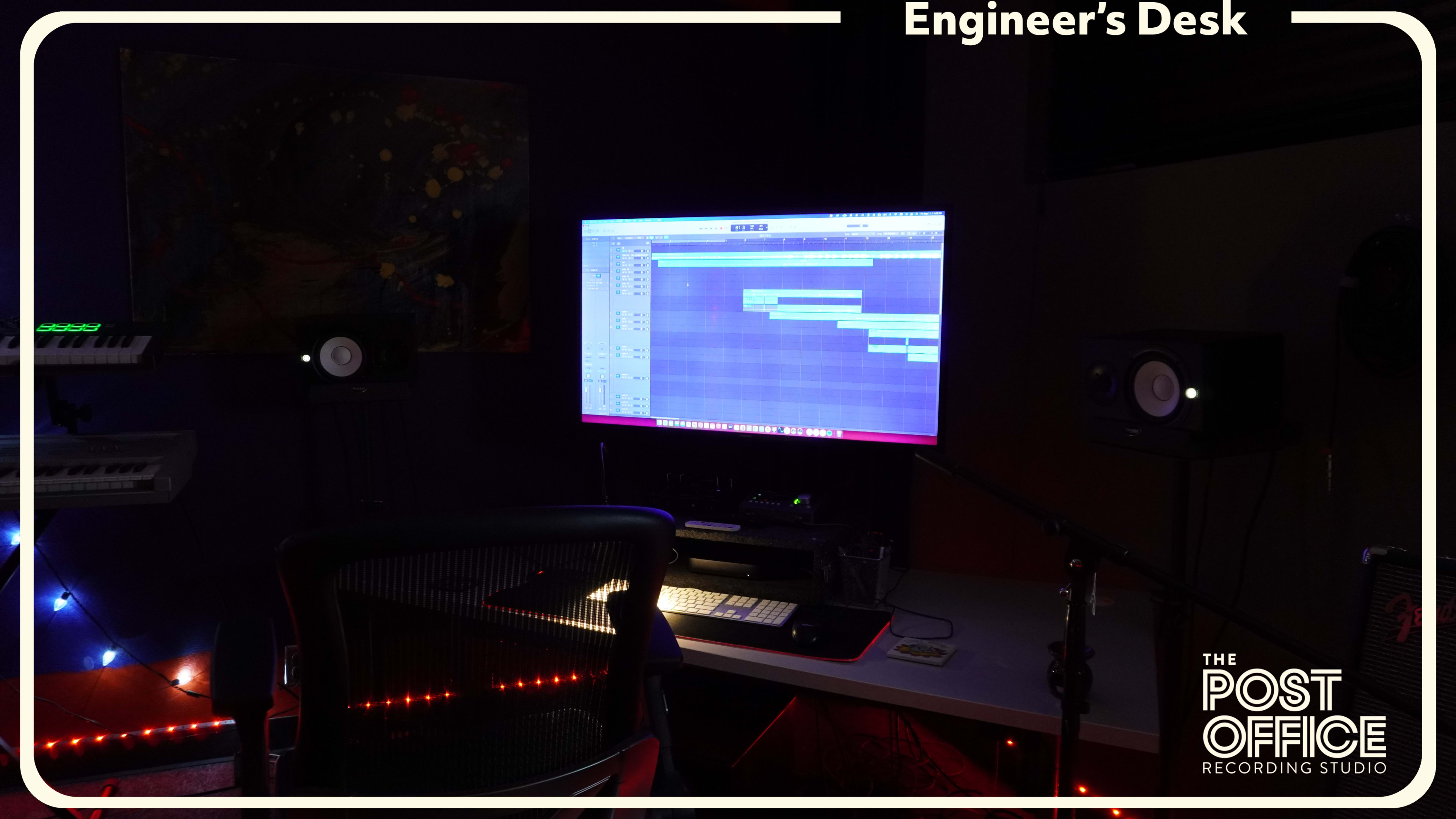 The image shows an engineer’s desk in a recording studio with a computer monitor displaying audio software and studio monitors positioned on either side.