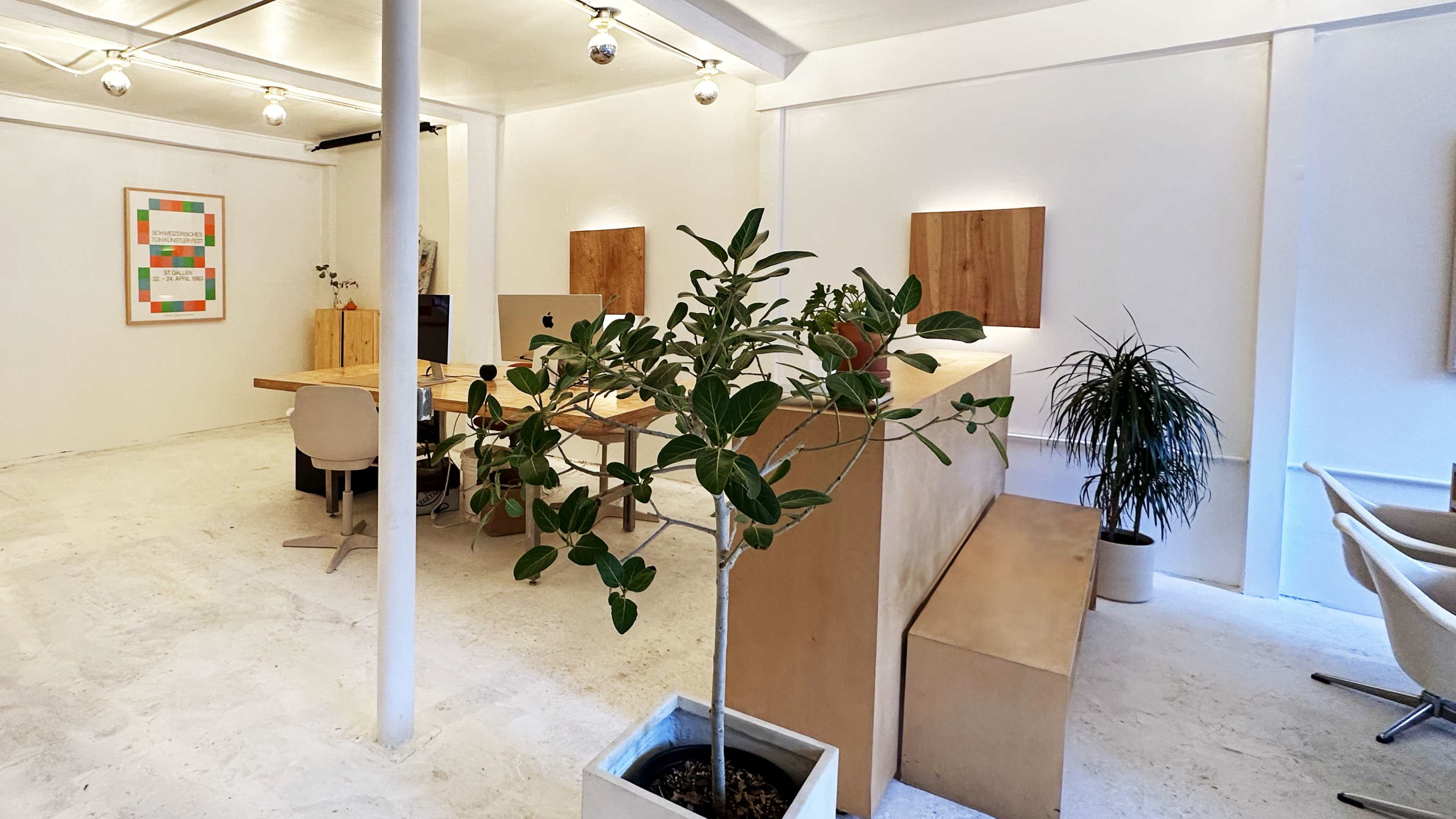 A modern office space features a central wooden table surrounded by chairs, potted plants, and artwork on the walls, with bright lighting and a minimalist aesthetic.