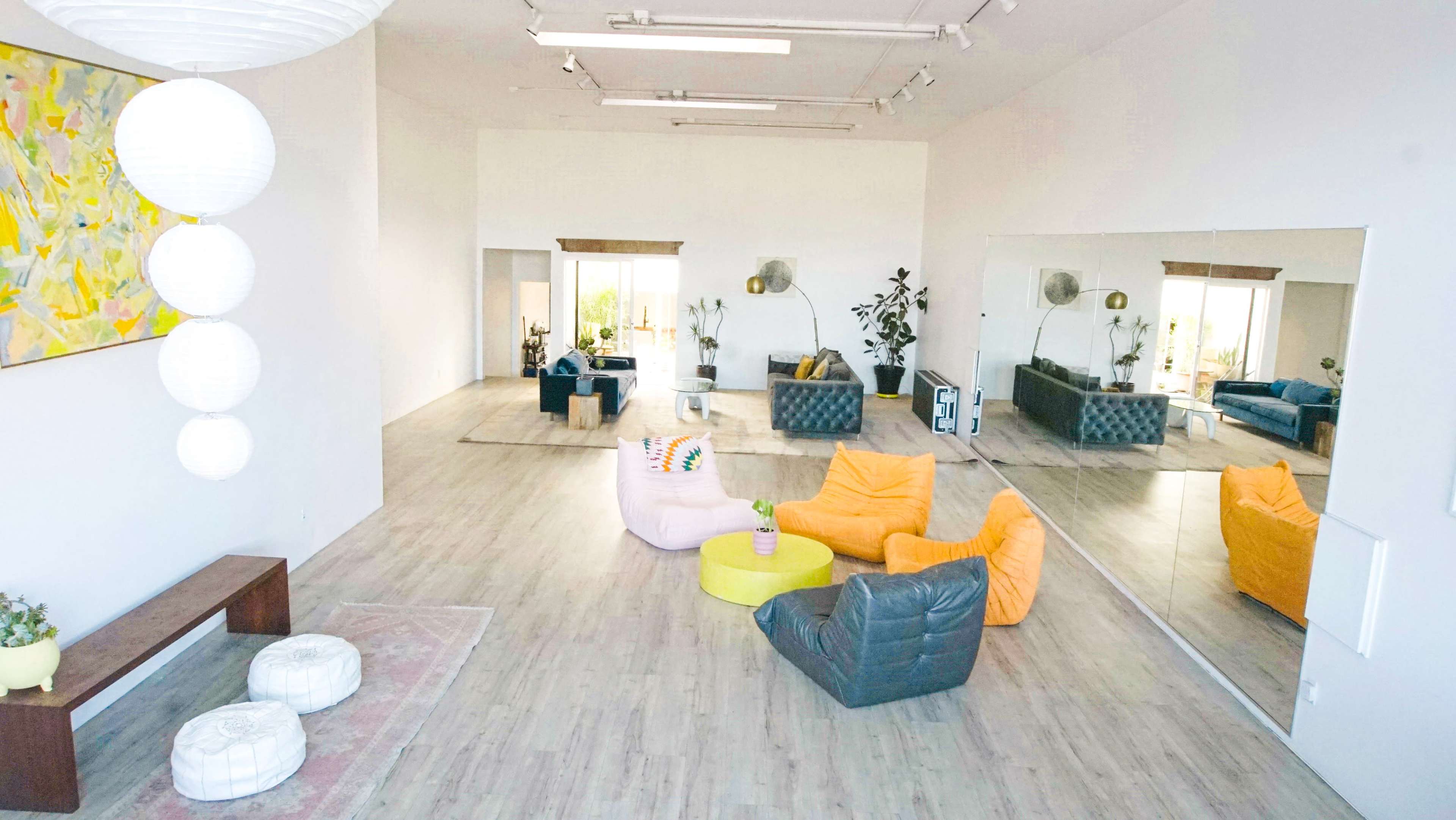 The image shows a spacious, well-lit interior with modern furniture, including colorful seating and a large mirror reflecting the room.