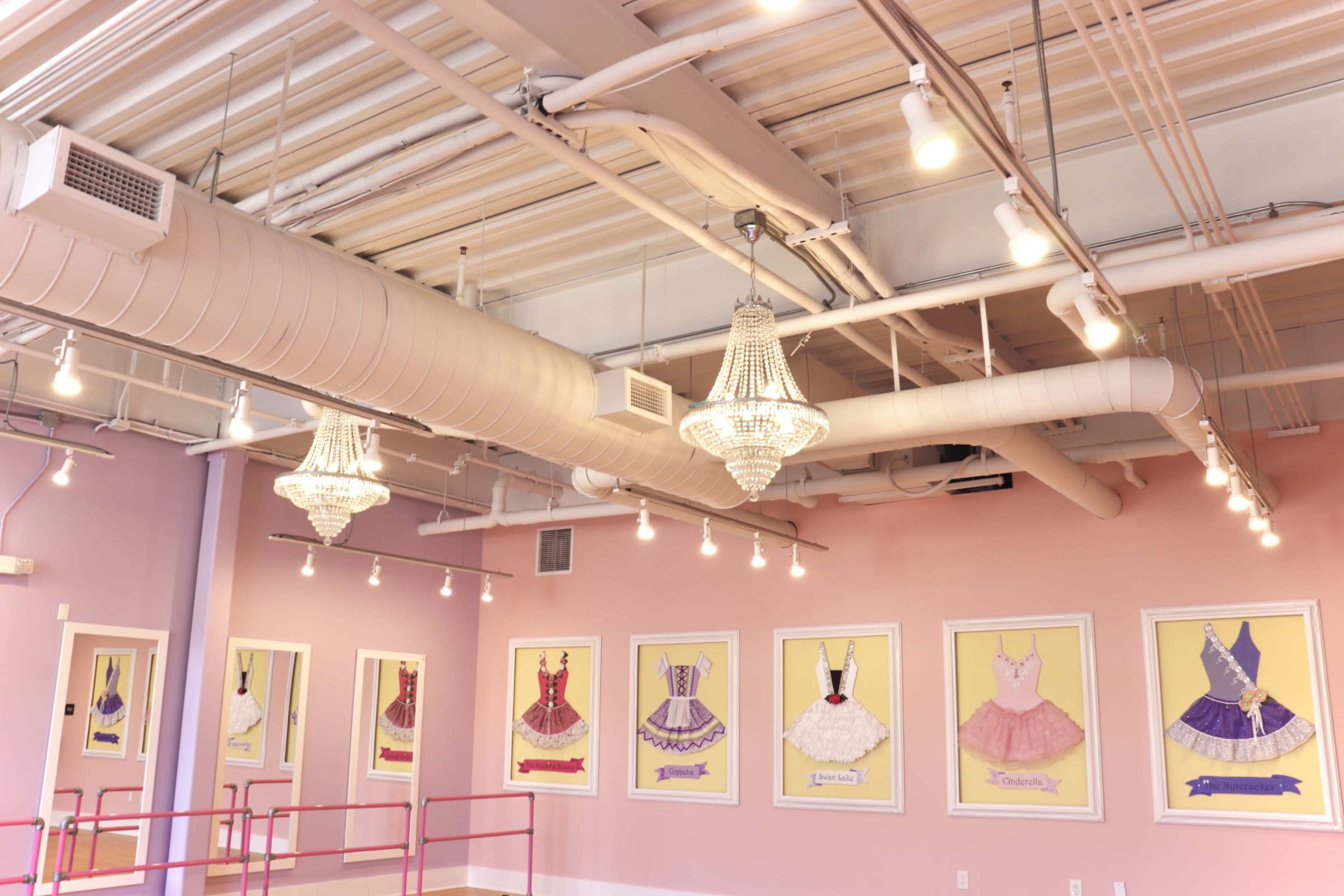 The image shows an interior of a room with a pink wall, chandeliers, and framed illustrations of dresses displayed along the wall.
