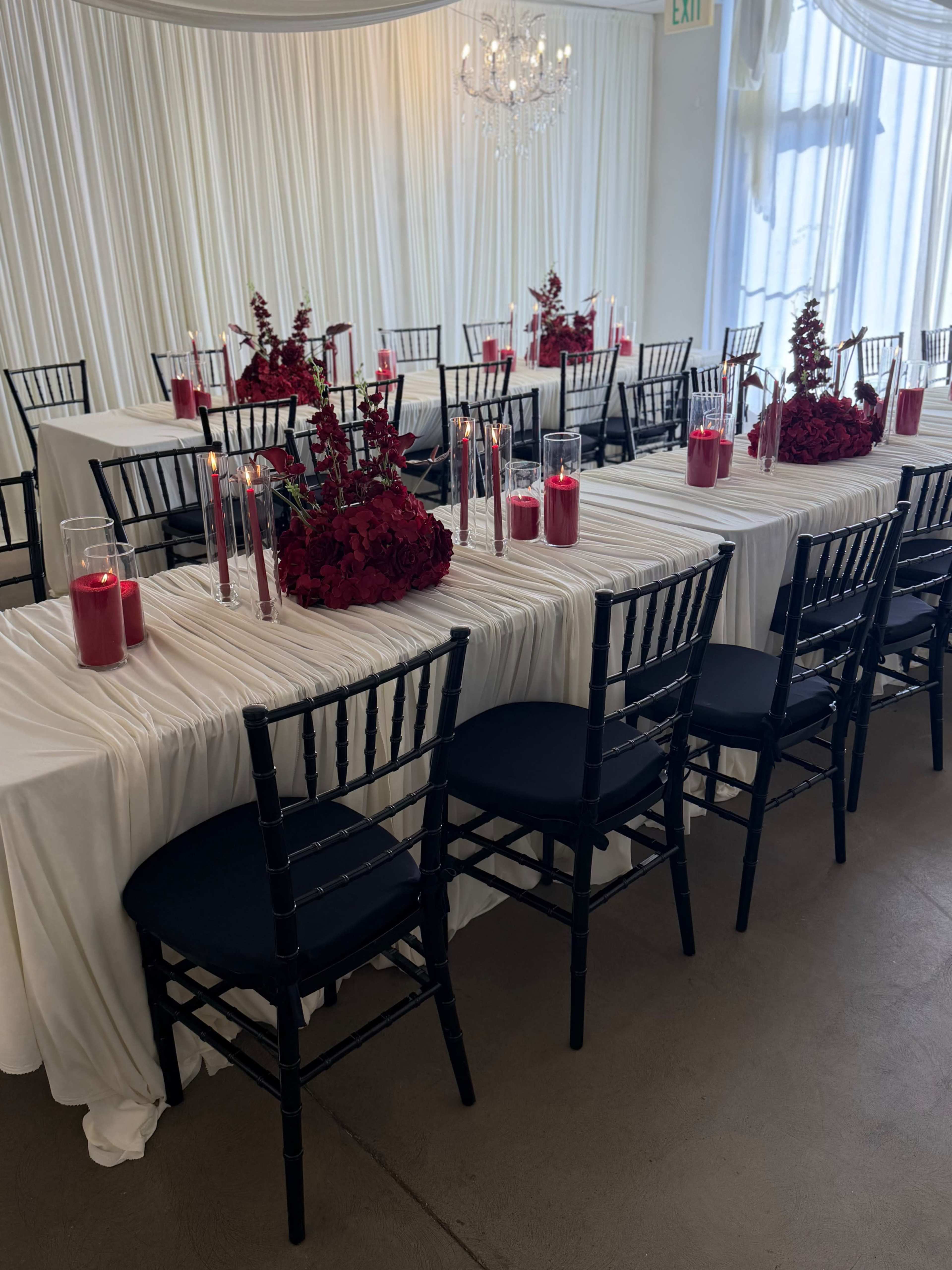 A banquet table adorned with white tablecloths, red floral arrangements, and candles is set up in a well-lit venue with a chandelier.