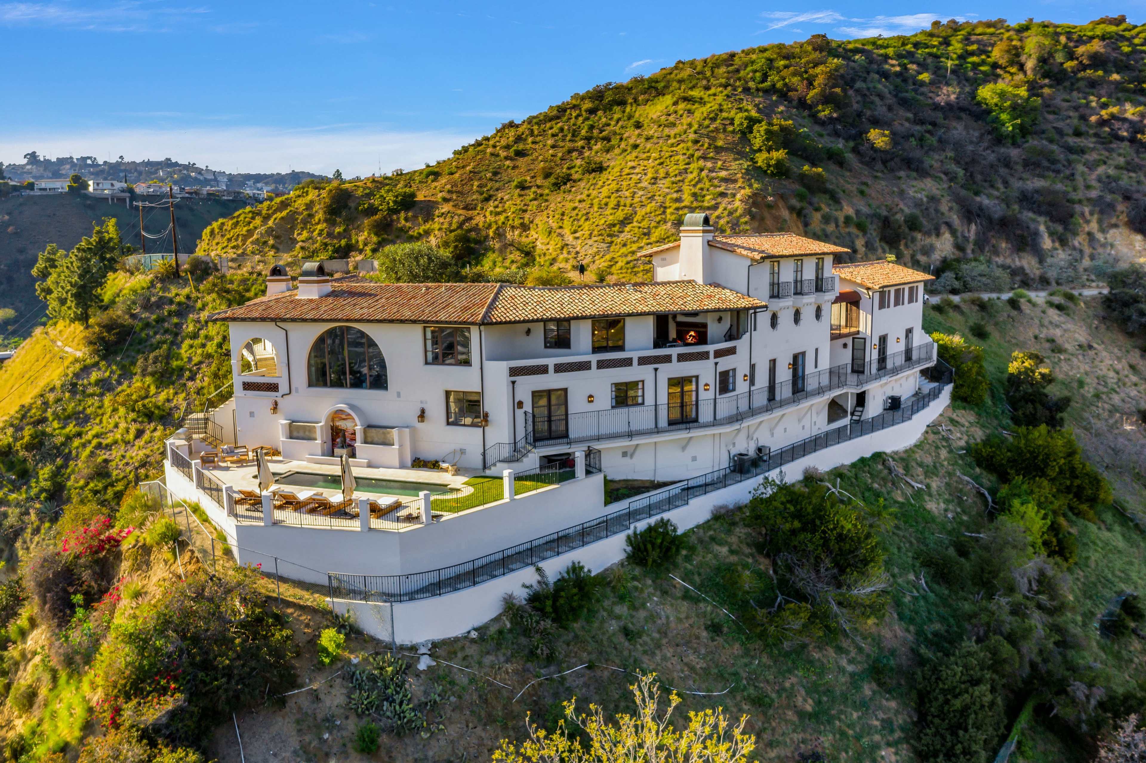 A large hillside mansion features multiple balconies and a swimming pool, surrounded by greenery.