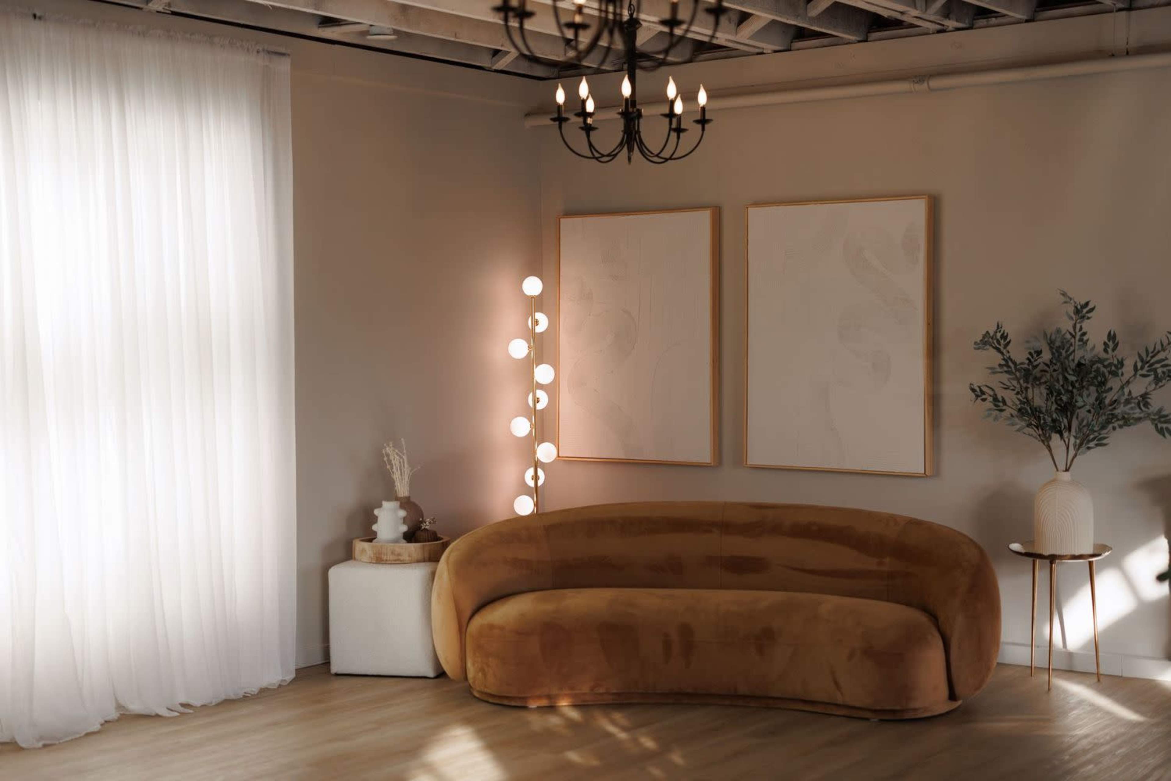 The image depicts a modern living space with a curved brown sofa, a minimalist white lamp, framed artwork on the wall, a potted plant, and natural light streaming through sheer curtains.