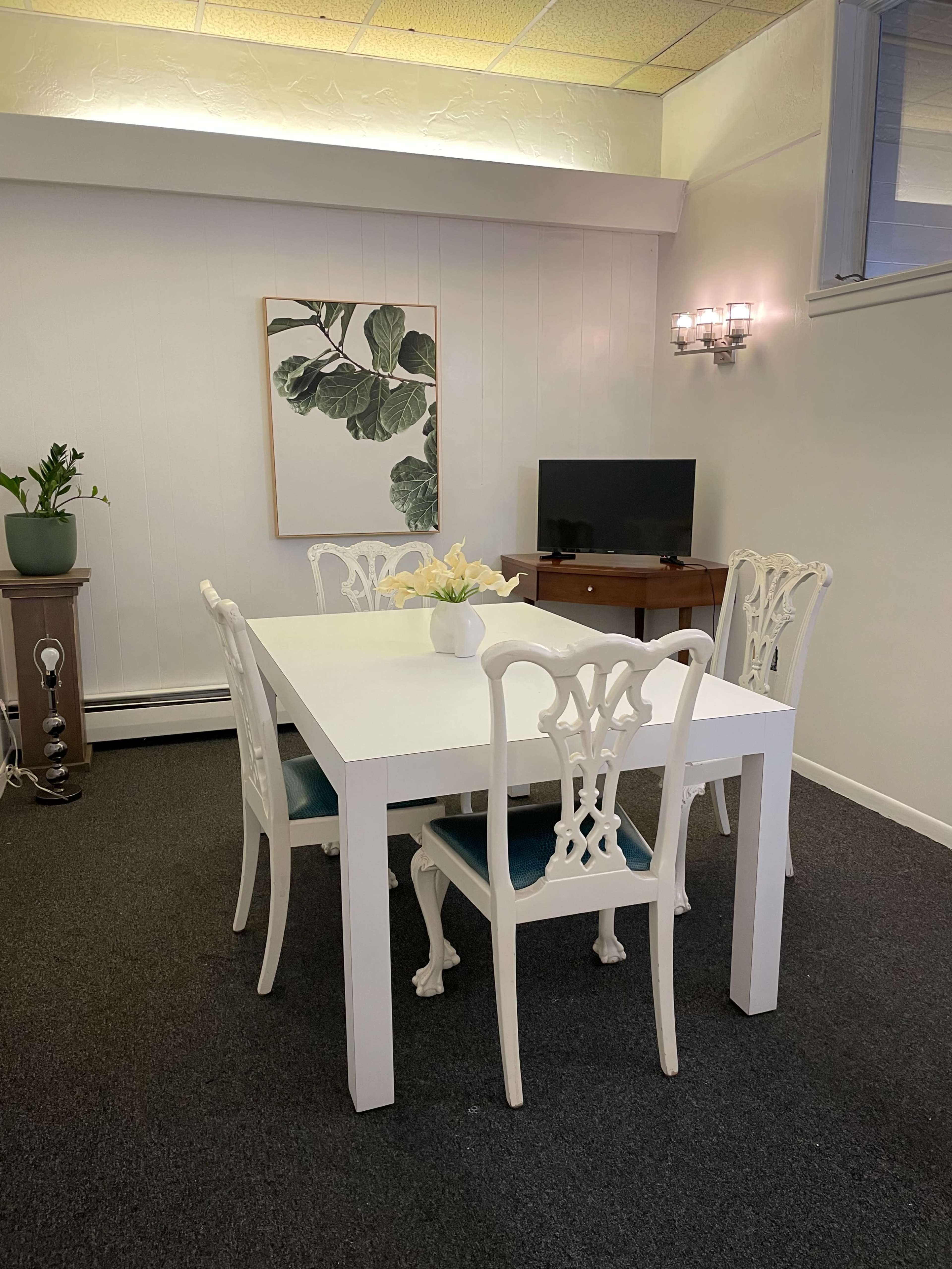 A white dining table with four chairs is set in a room featuring a plant, a wall-mounted television, and a piece of artwork.