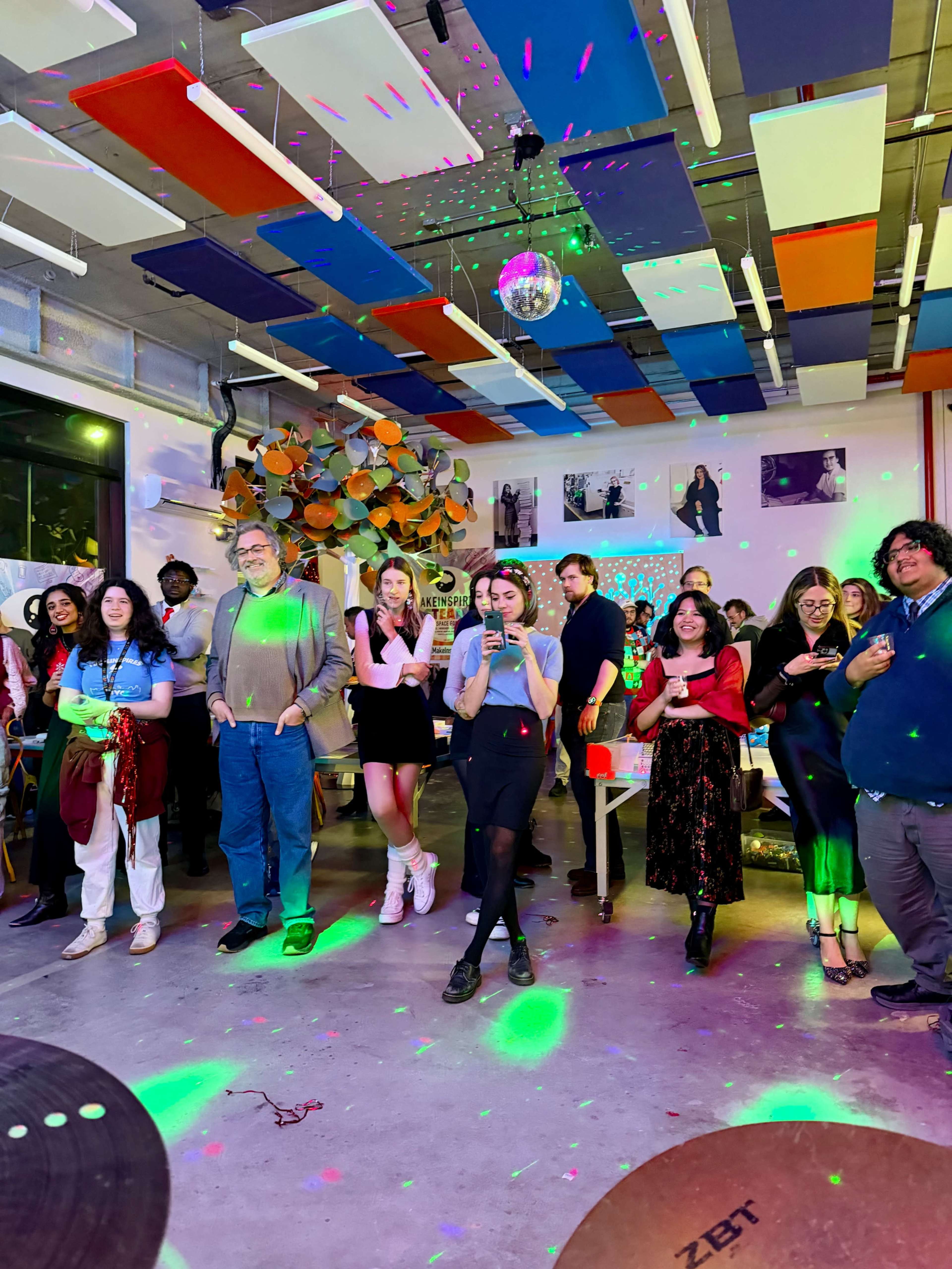 A group of people stands together in a colorful, lively indoor space decorated with art, while disco lights reflect off a mirror ball above them.