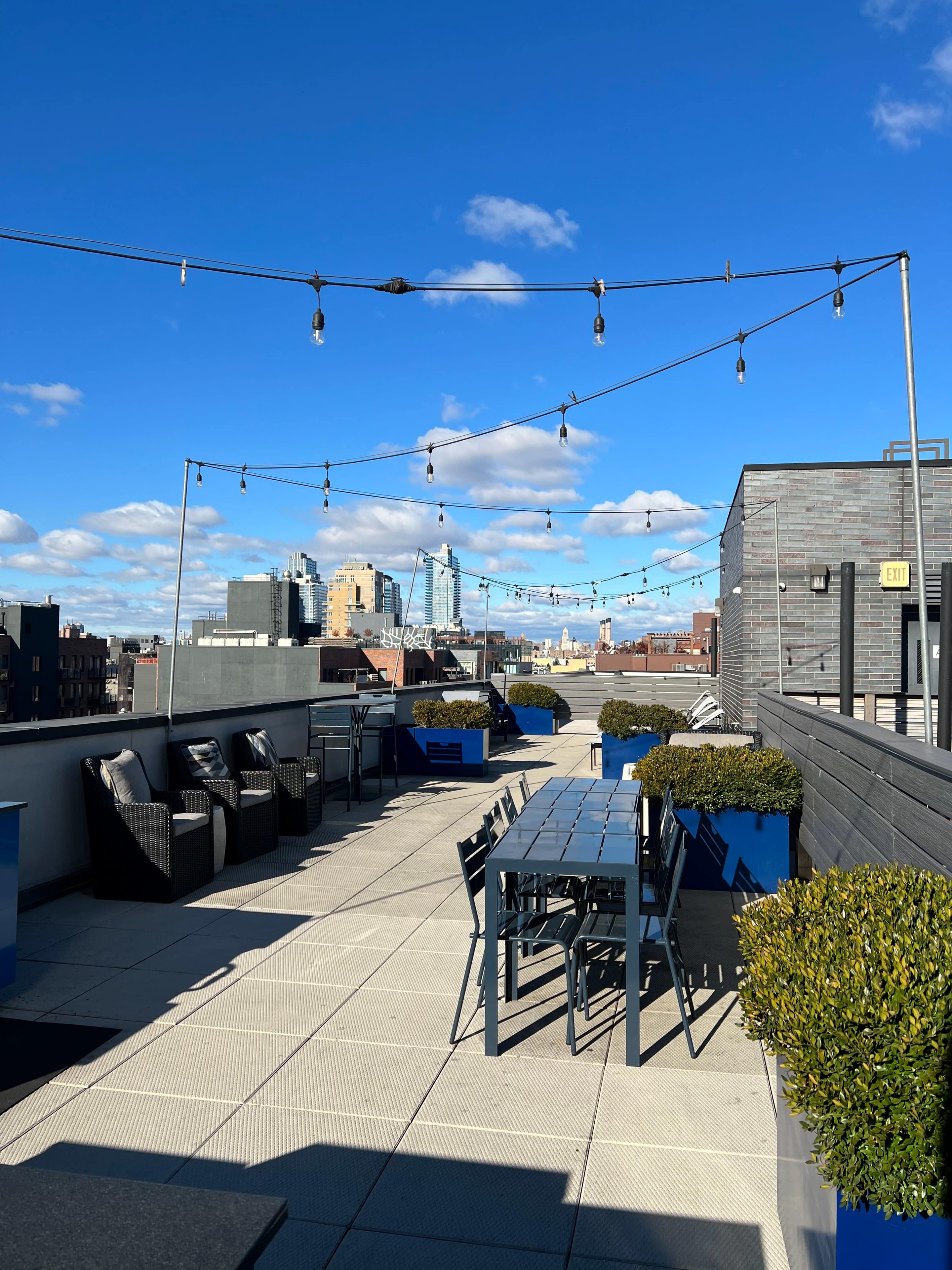 A rooftop terrace with seating areas, a dining table, and decorative string lights against a clear blue sky.