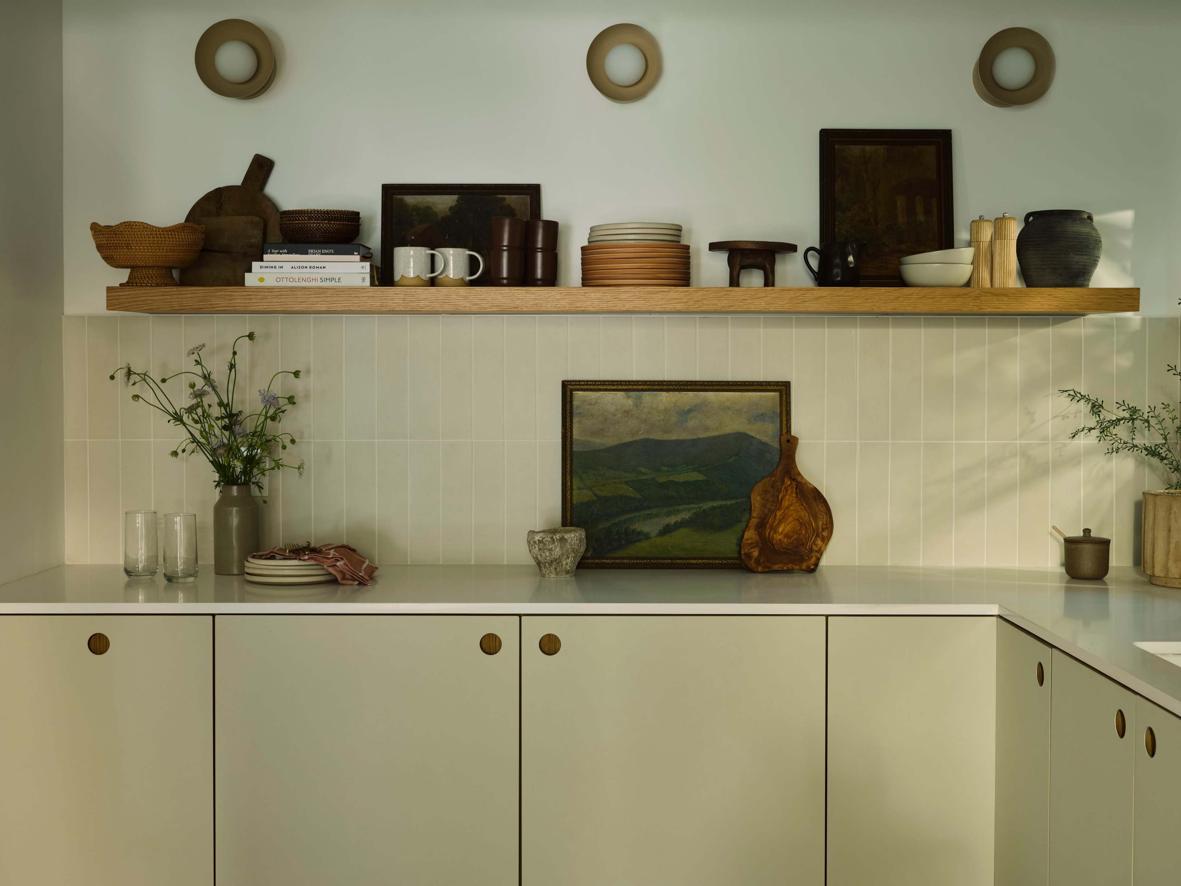 A modern kitchen features a shelf with various dishes, decorative items, and a landscape painting above streamlined cabinetry.