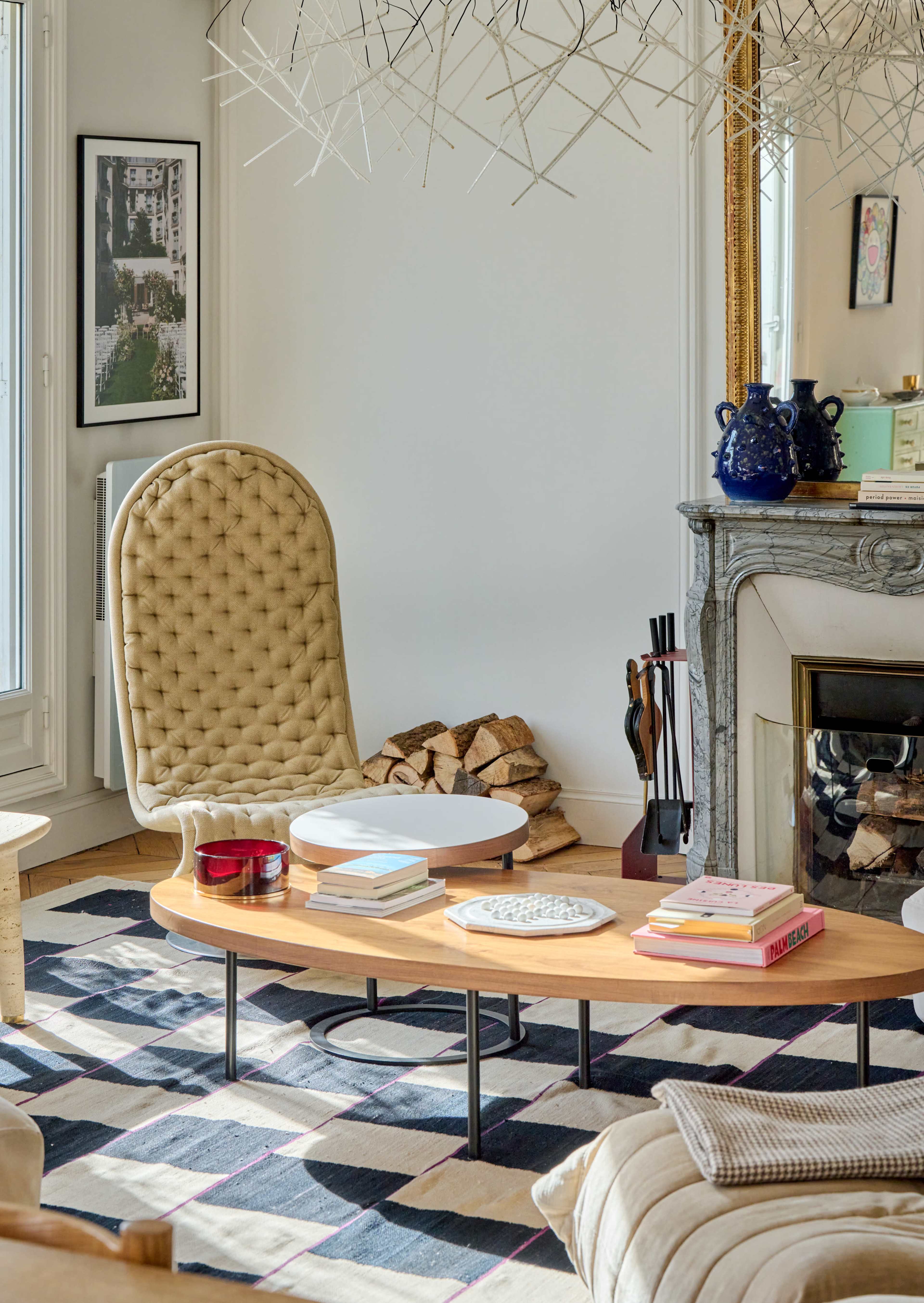 A modern living room featuring a tufted beige high-back chair, a coffee table with various books and decorative items, and a fireplace adorned with a mirror and styled accessories.