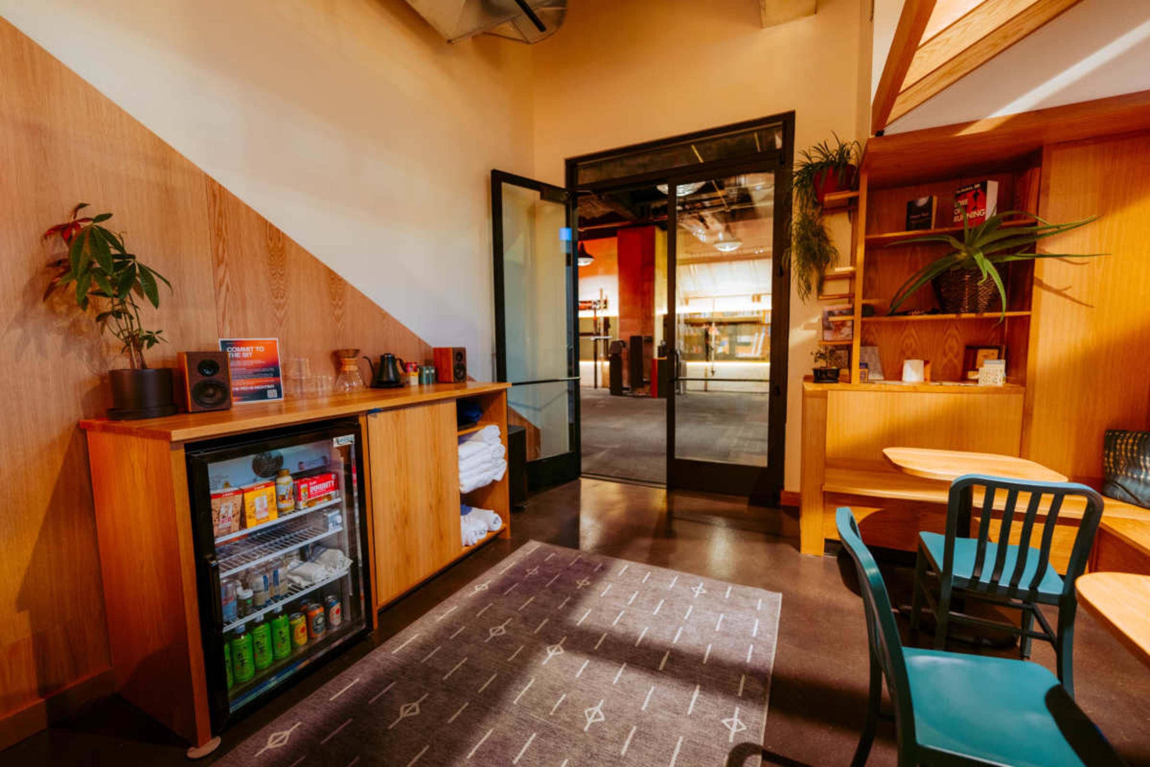 The image shows a modern cafe interior with wooden furnishings, a small refrigerator filled with beverages, and a view of an open space through glass doors.