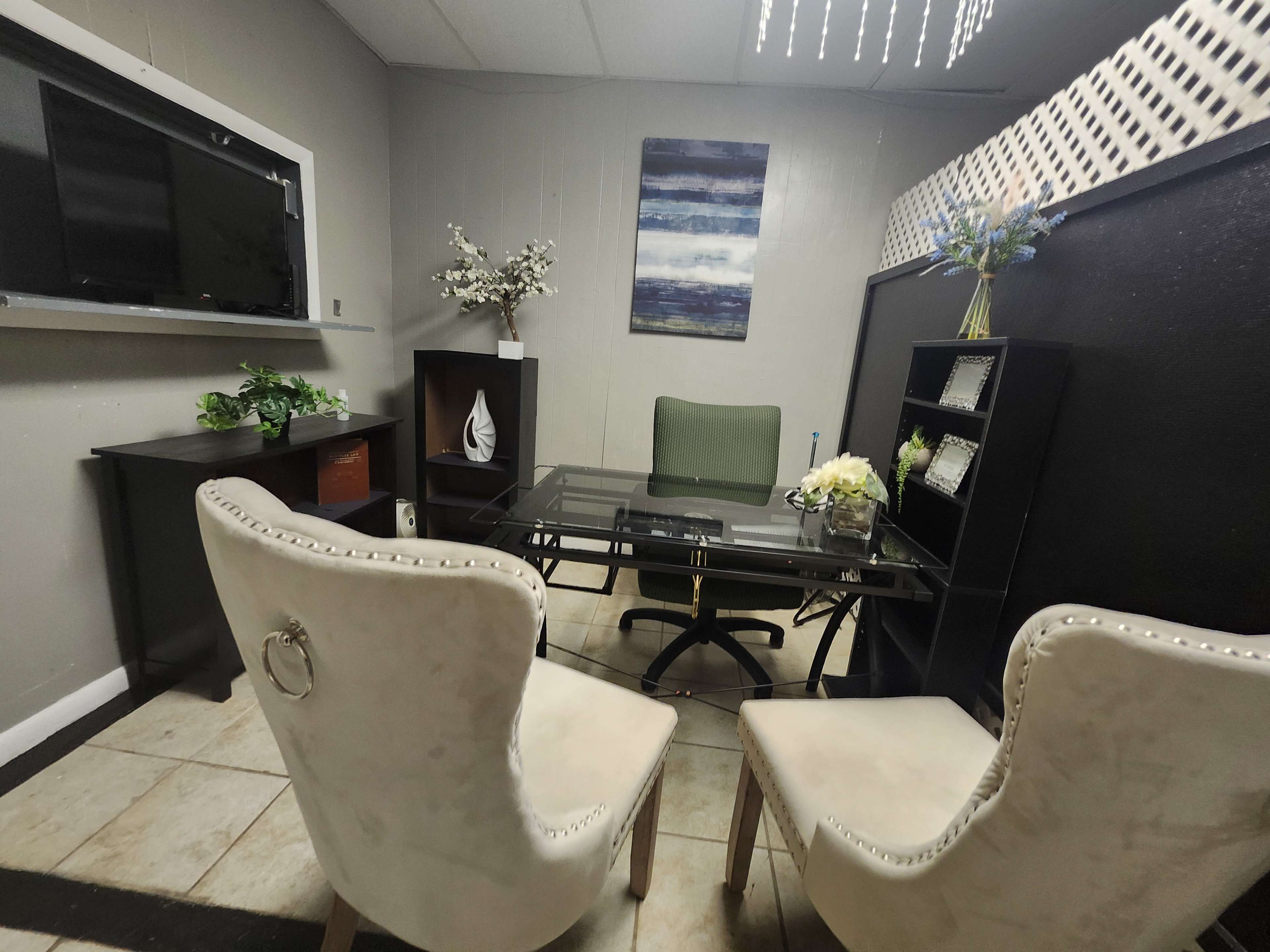 The image shows a small office space featuring a glass-top desk, two upholstered chairs, a decorative wall shelf, and a television mounted on the wall.