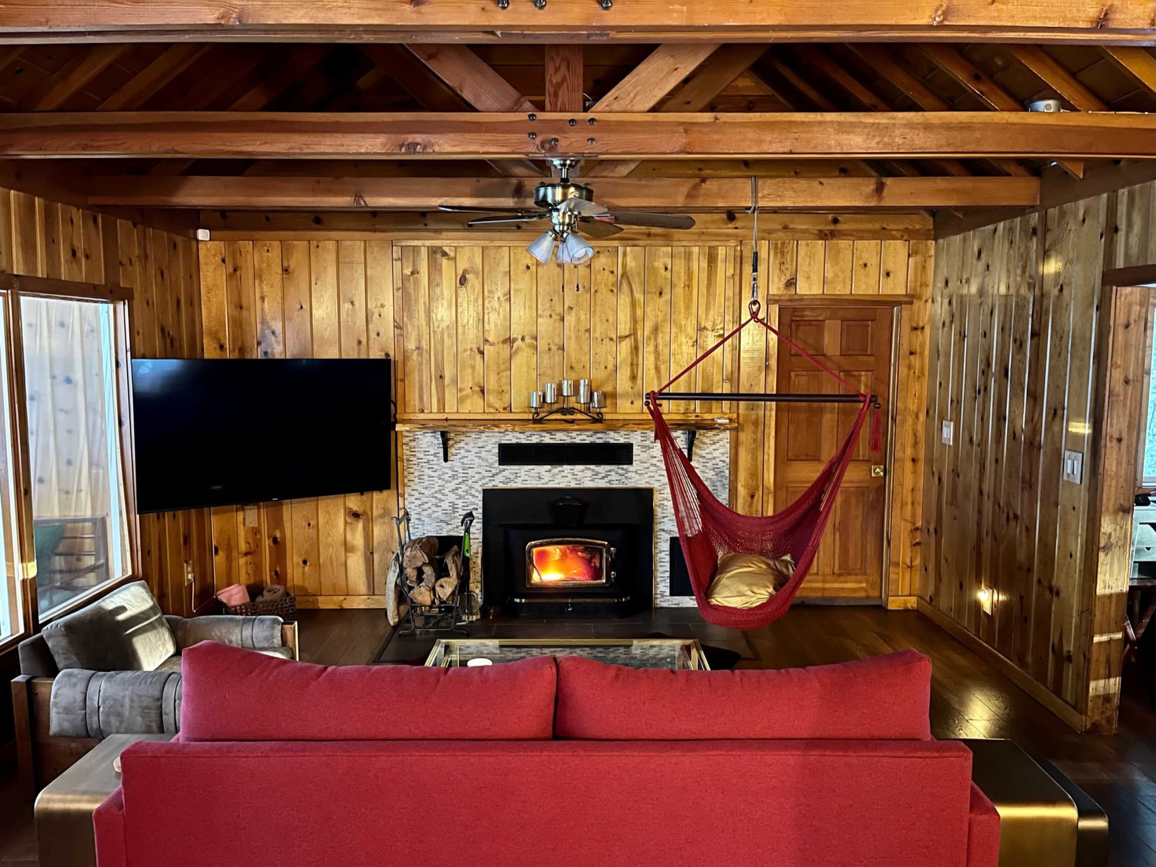 A wooden cabin interior featuring a red sofa, a television, a fireplace with a fire, and a hammock hanging from the ceiling.