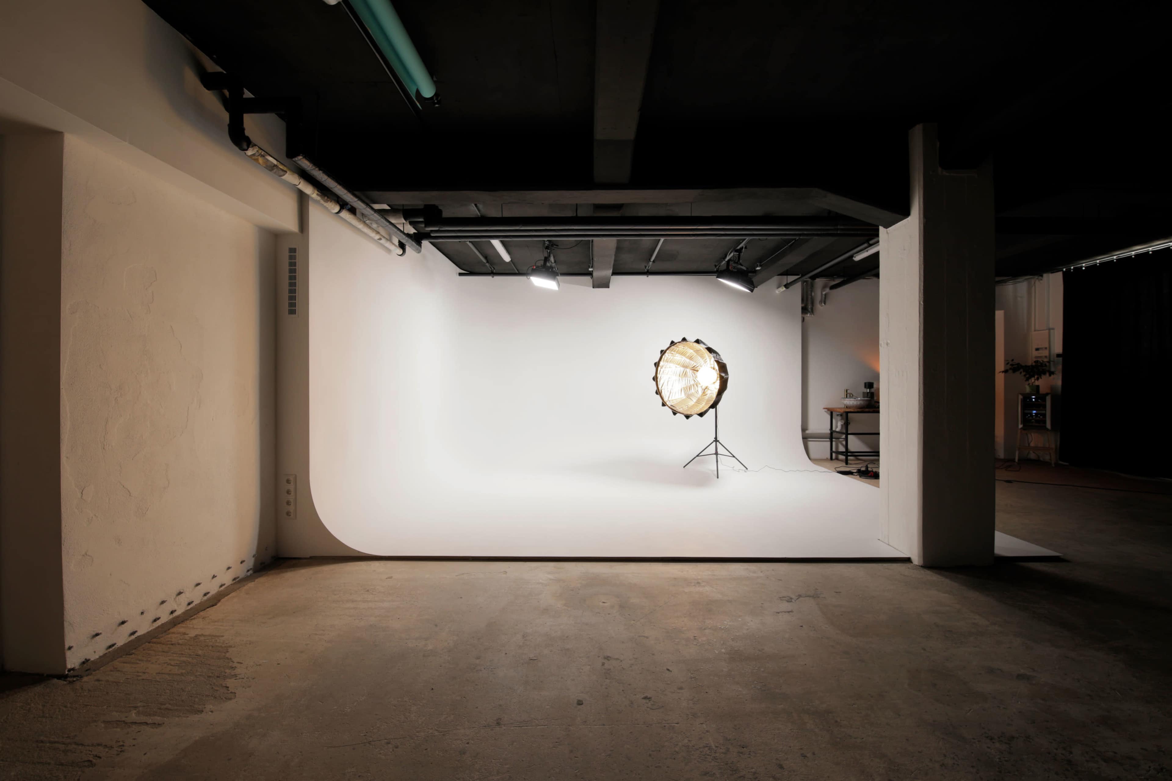 A photography studio features a circular light source on a stand in front of a white backdrop, surrounded by concrete walls and a dark ceiling.