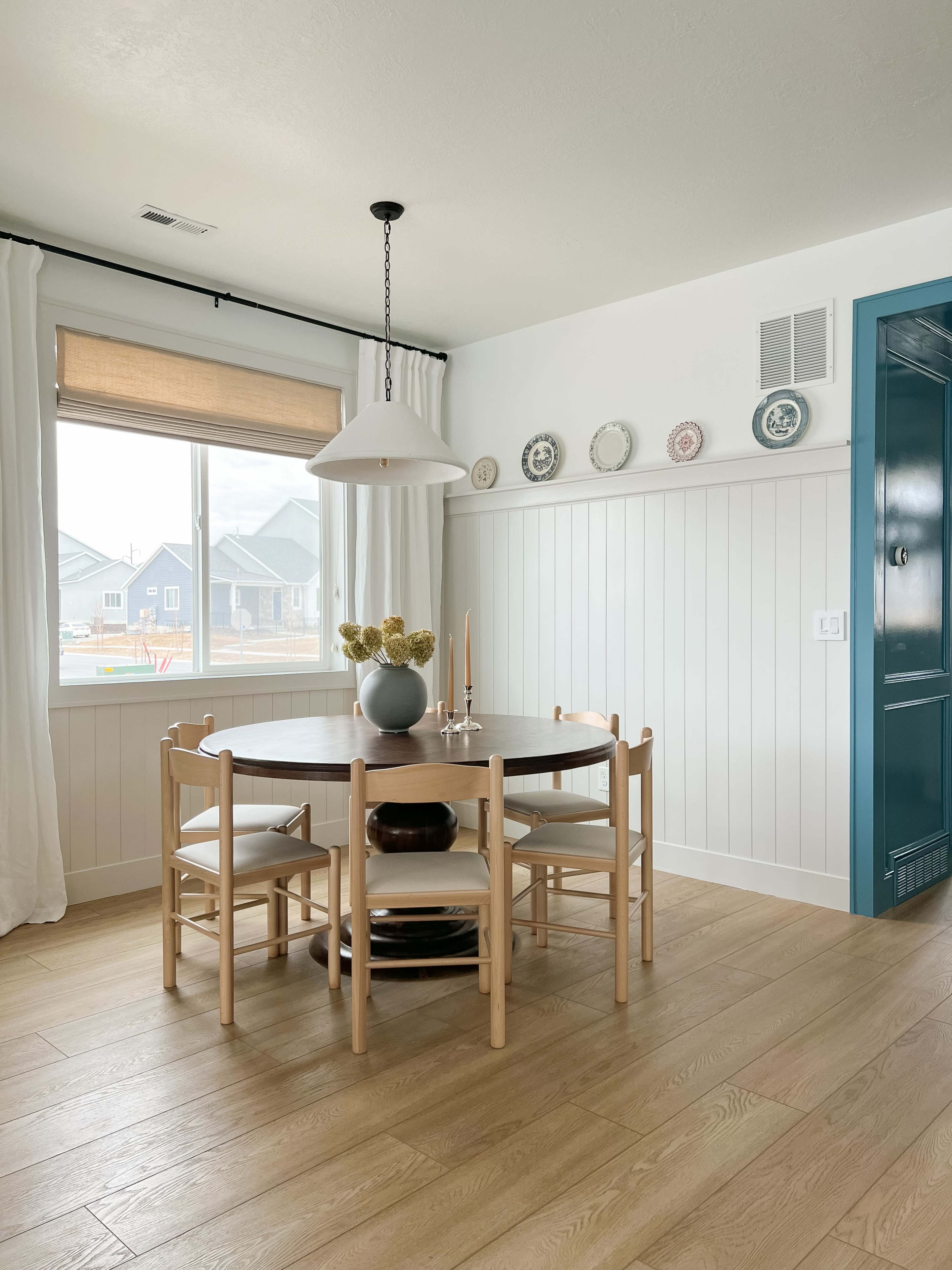 A round wooden dining table surrounded by chairs, with a large window and decorative plates on the wall in a bright, minimalistic dining area.