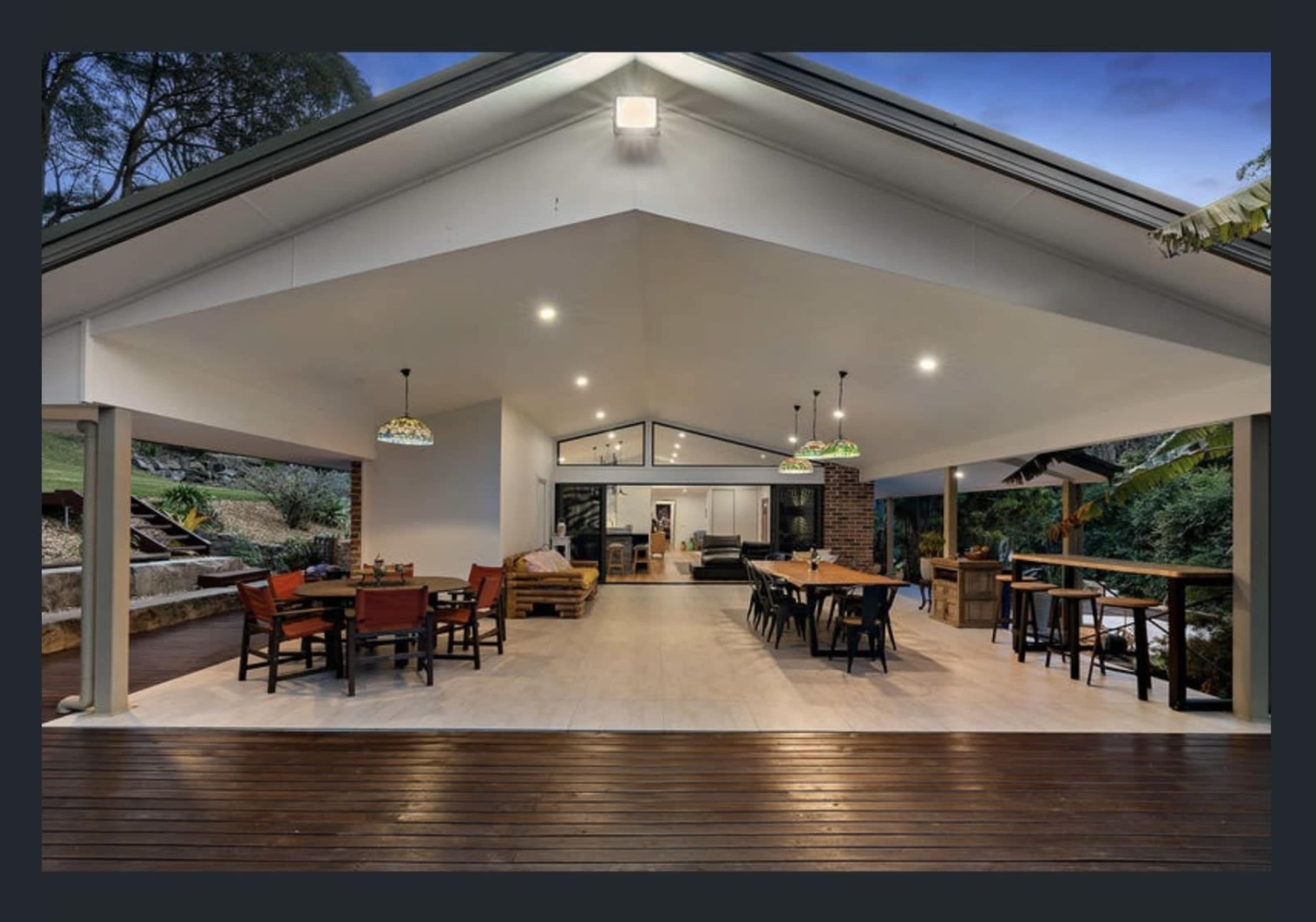 A spacious outdoor living area with wooden flooring, featuring dining tables and chairs under pendant lights, connected to a modern interior space.