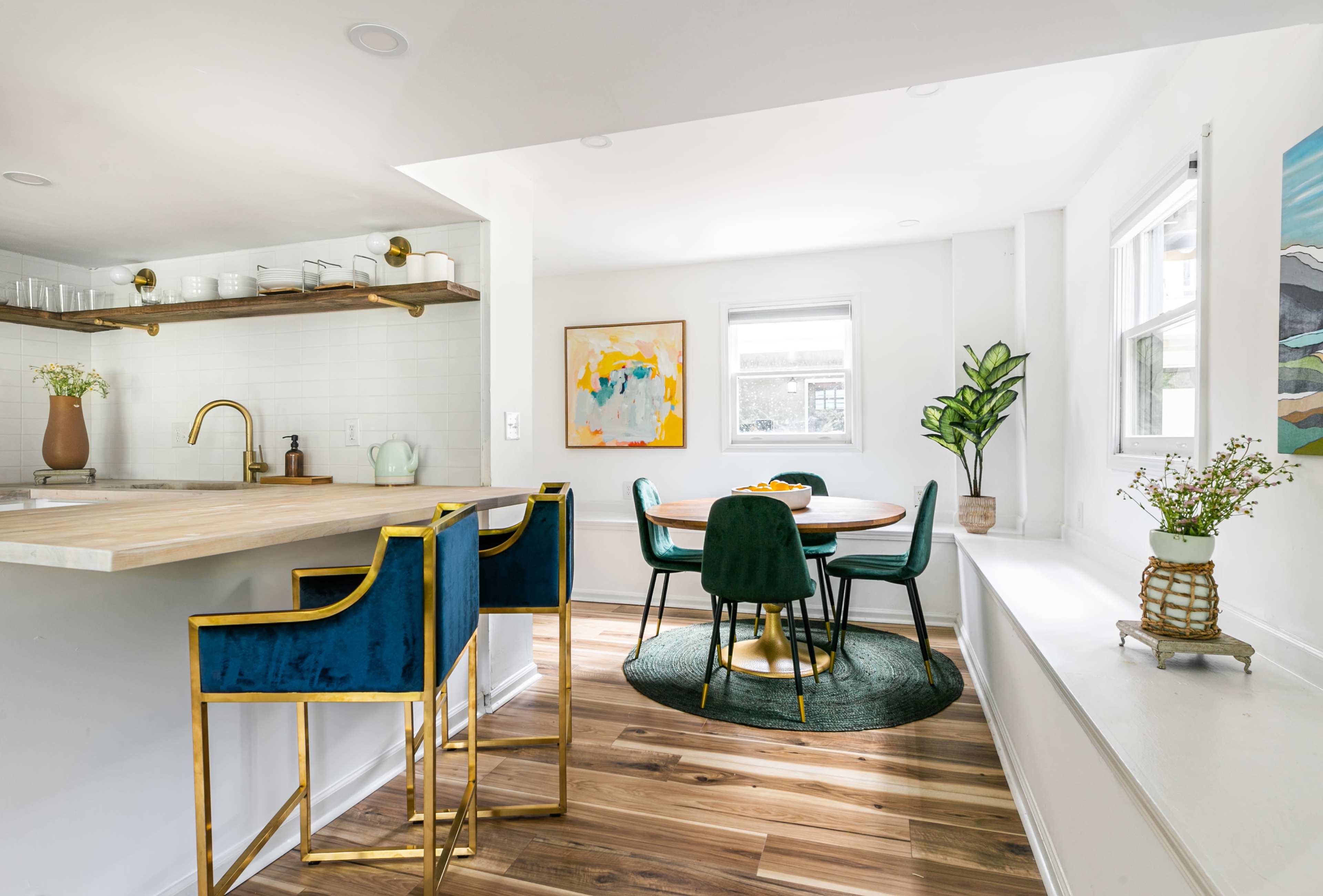 A modern kitchen with a wooden island and gold bar stools, adjacent to a dining area featuring a round table surrounded by chairs and a potted plant.