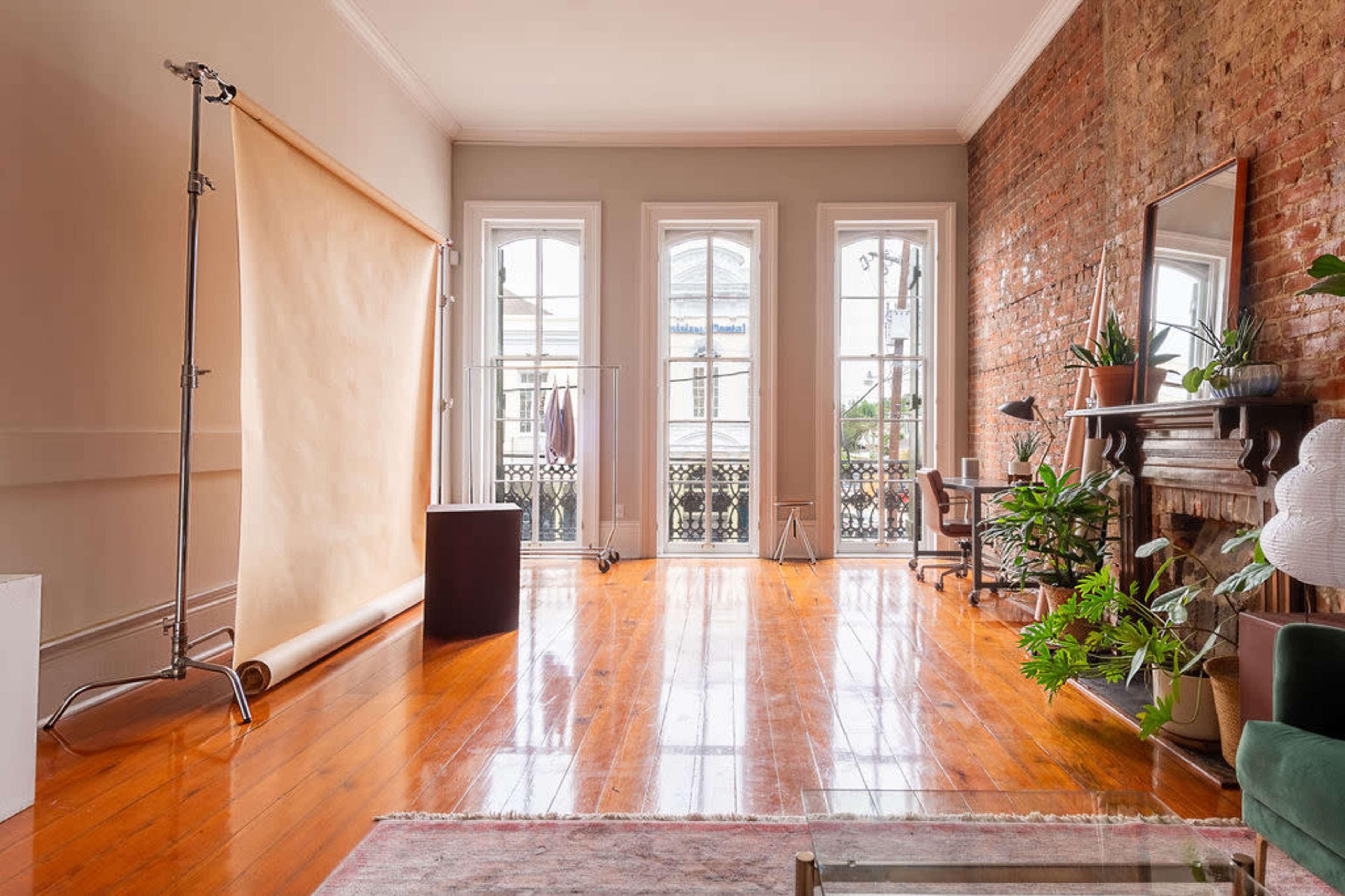 A spacious room with large windows, hardwood floors, a brick wall, a backdrop stand, and a mirror.