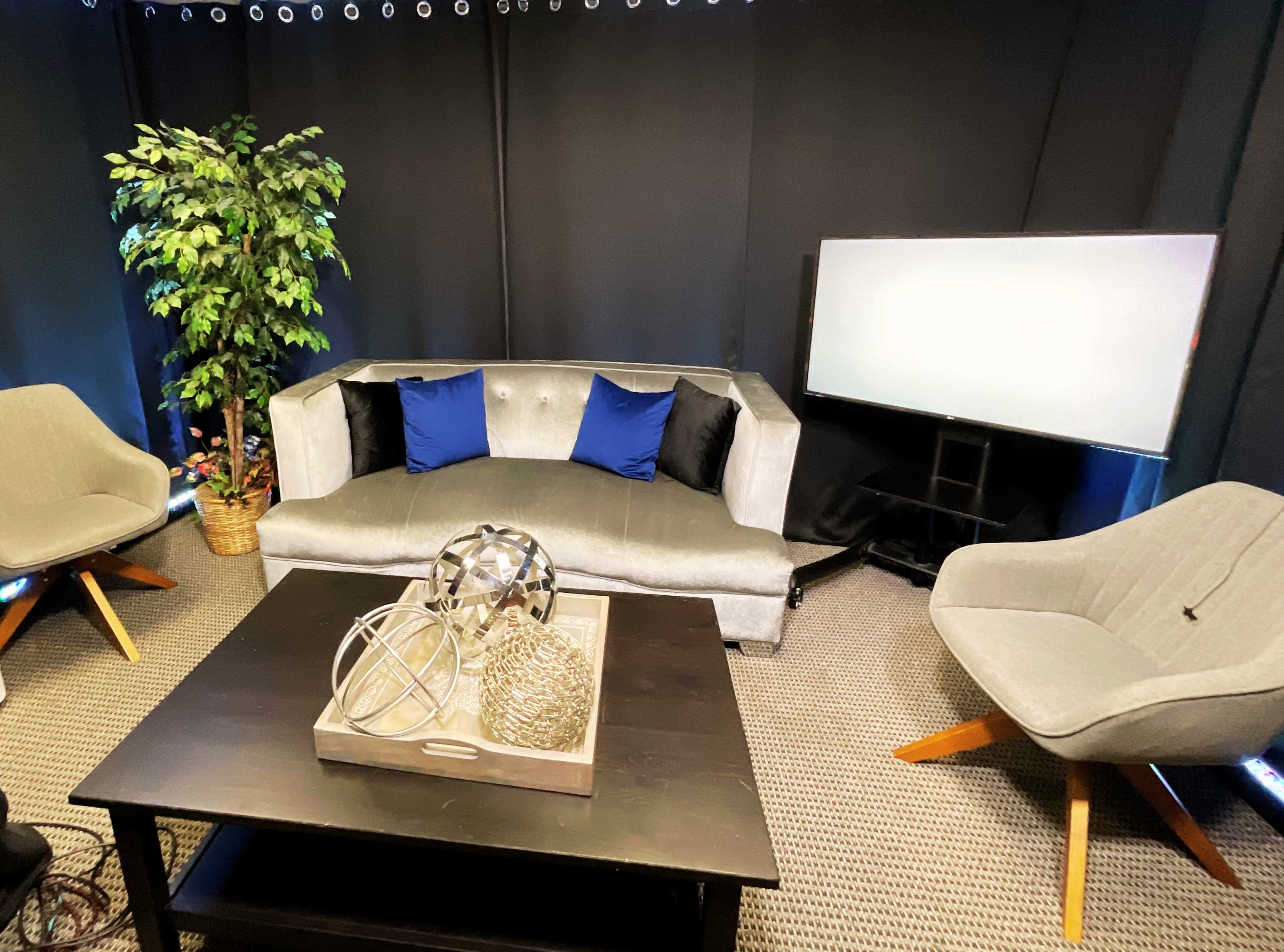 The image shows a modern seating area with a gray couch adorned with blue and black pillows, two gray armchairs, a black table with decorative items, and a blank TV screen against a black backdrop.