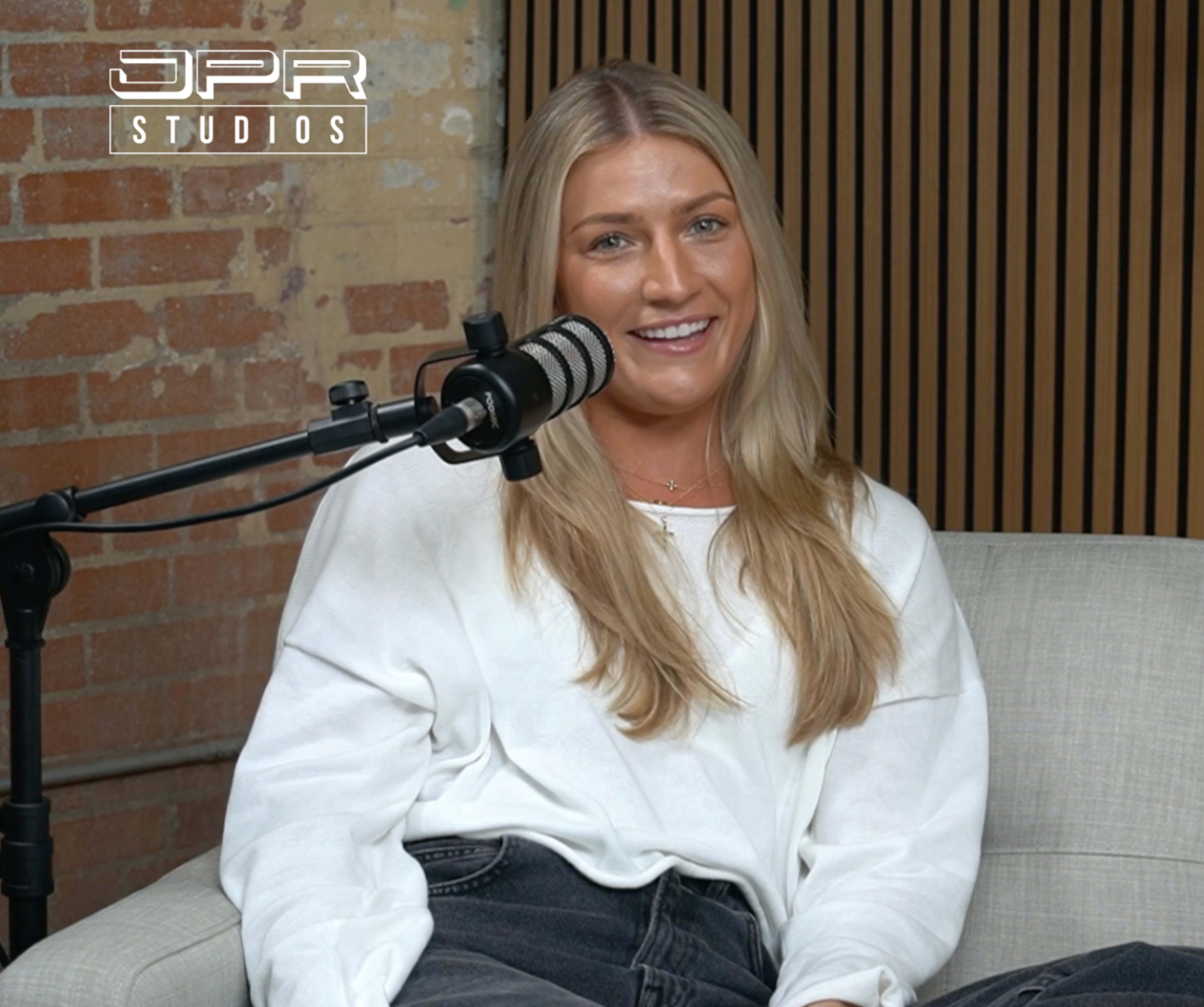 A woman with long blonde hair sits on a light-colored couch in a studio, smiling while speaking into a microphone.
