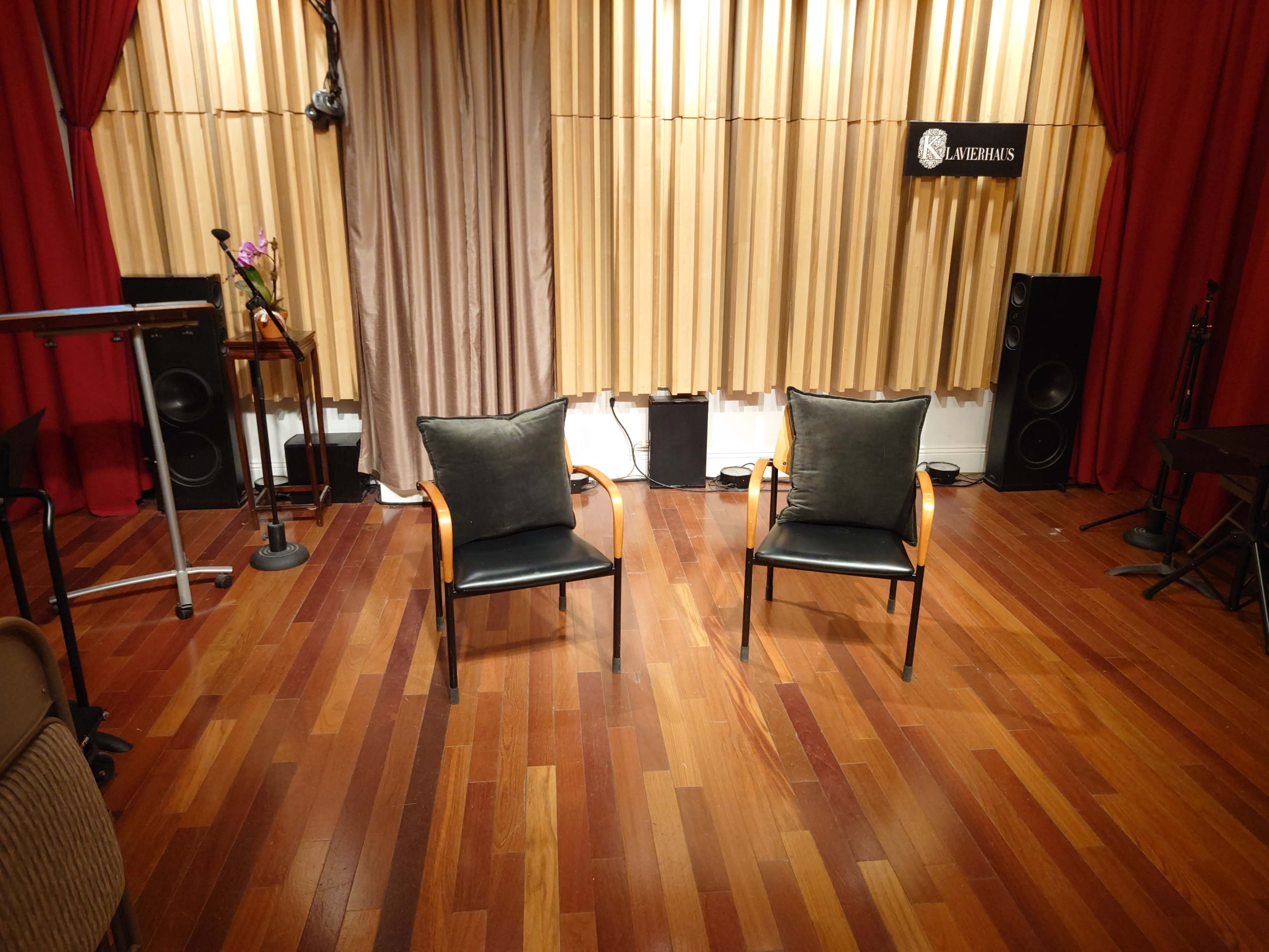 The image shows a room with two black chairs positioned facing each other on wooden flooring, flanked by curtains and audio equipment.
