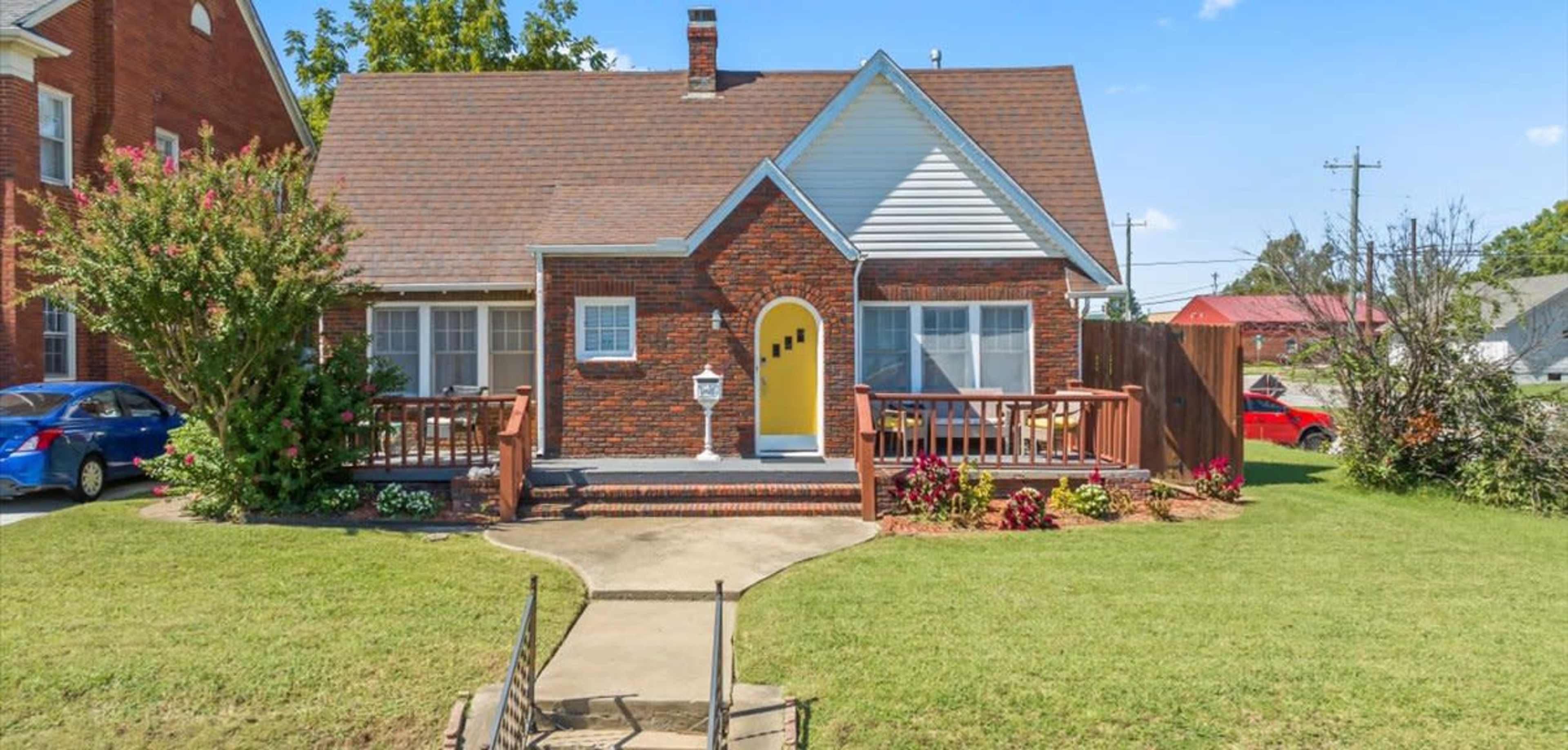 A brick house with a gabled roof features a front porch, colorful landscaping, and a small pathway leading to the entrance.